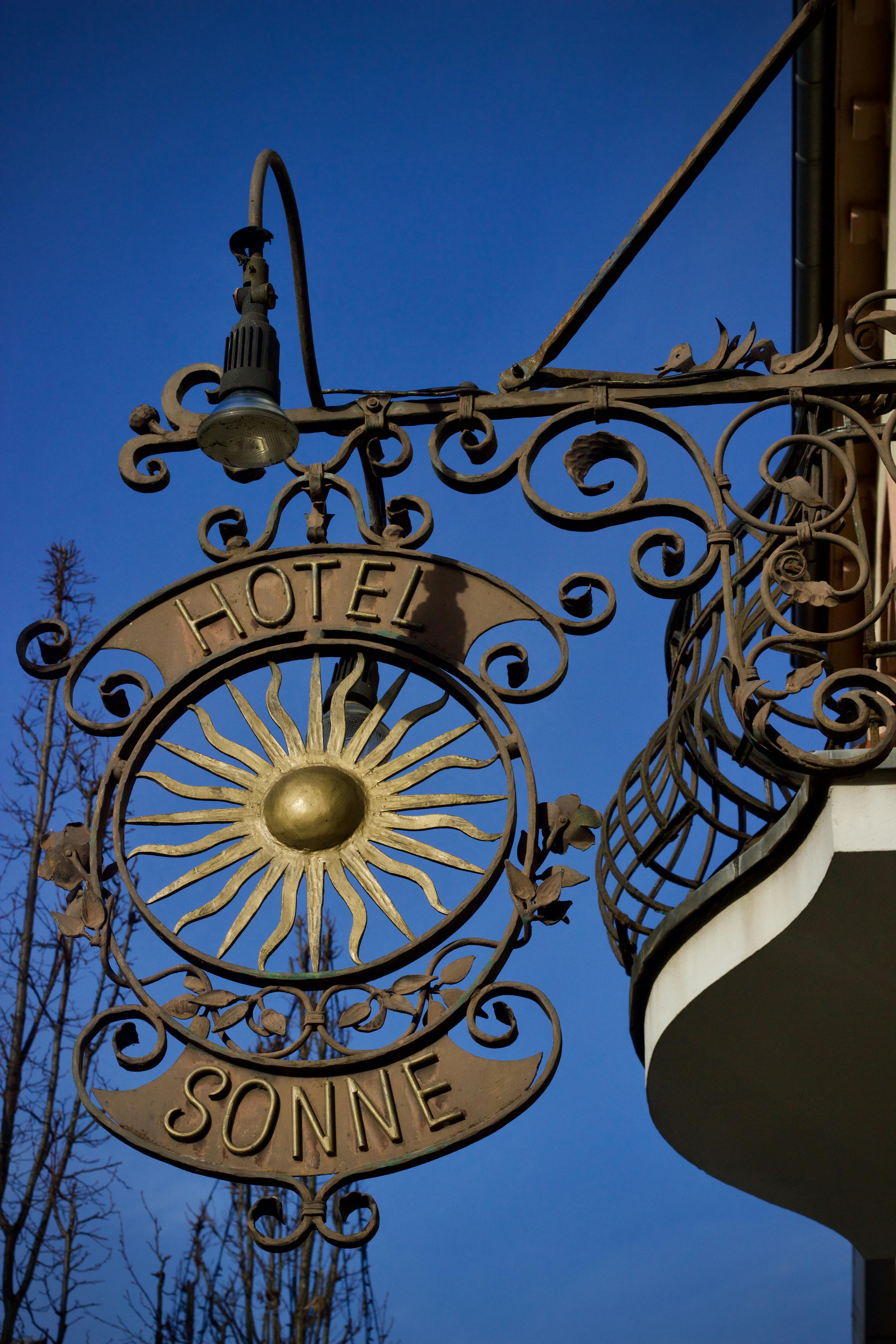 Gratis Papan nama Hotel Sonne dari logam artistik dengan lambang matahari di atas langit biru cerah. Foto Stok