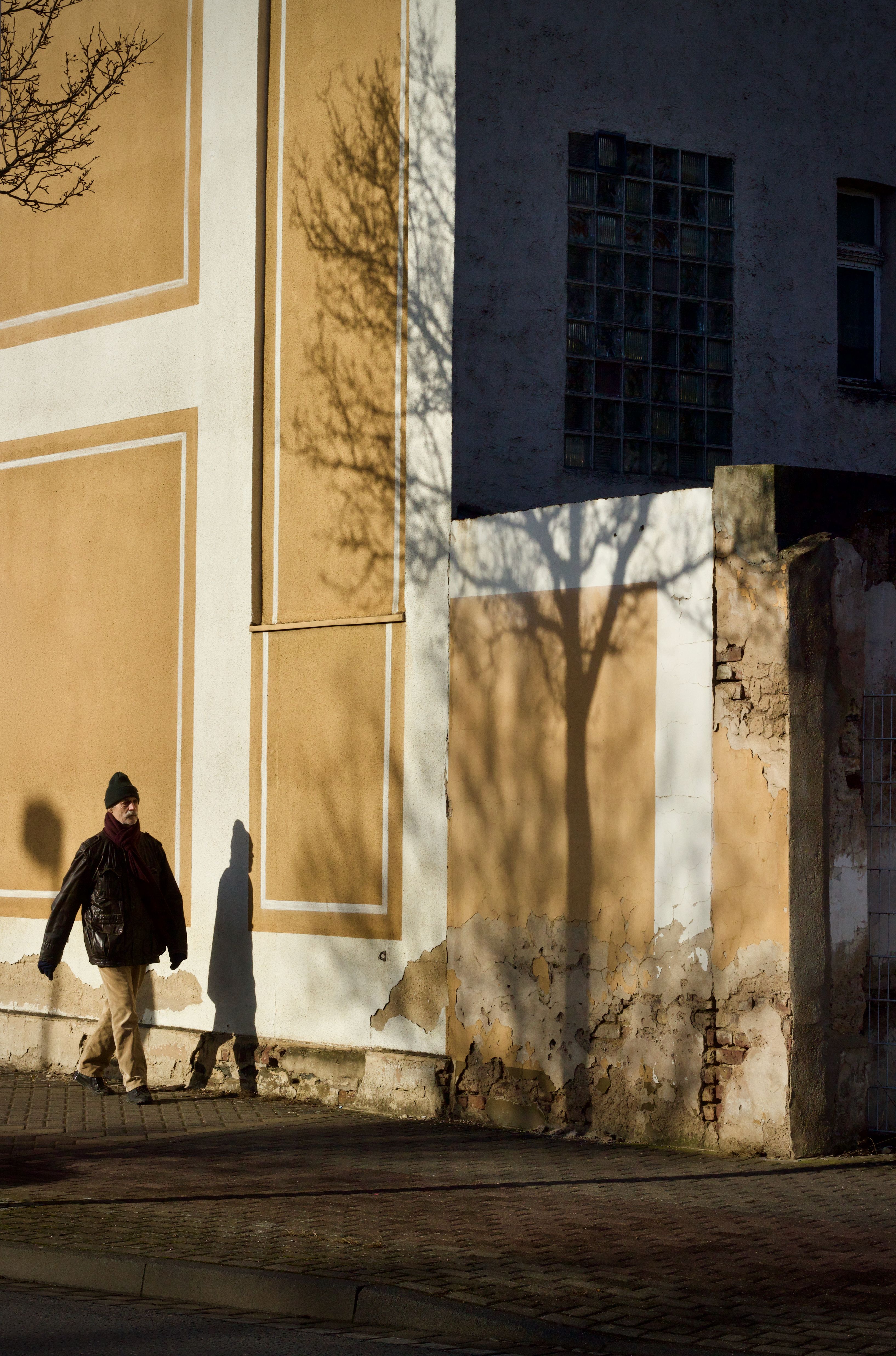 A man walks along a sunlit wall in an urban street, casting shadows.