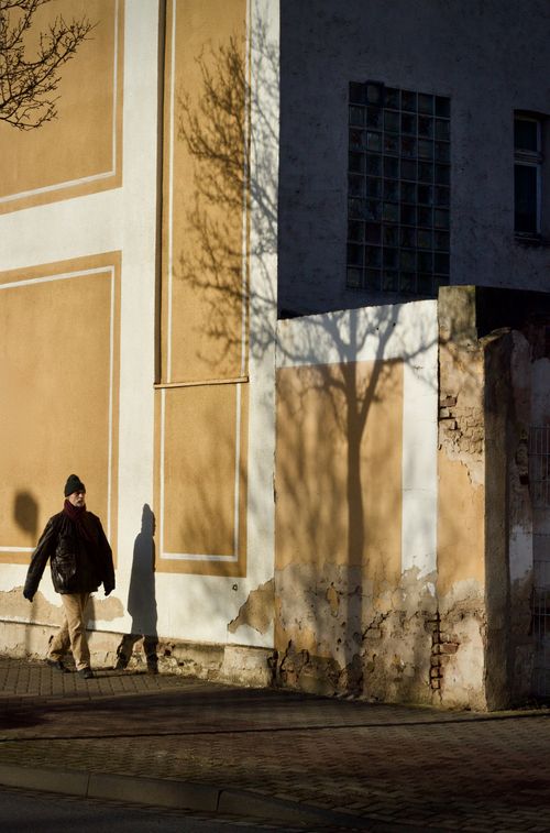 Free A man walks along a sunlit wall in an urban street, casting shadows. Stock Photo
