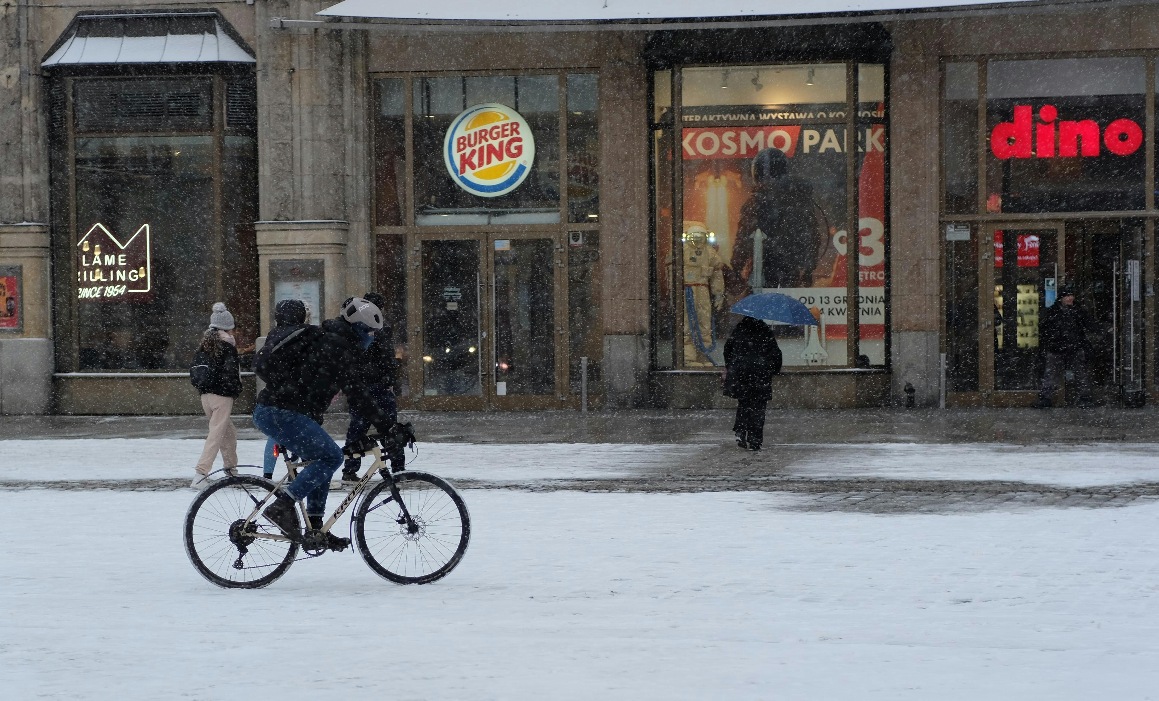 Cyclist rides through snowy city street with pedestrians and store signs.