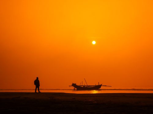 Free Silhouette of a person and boat on the beach under a vibrant orange sunset sky. Stock Photo