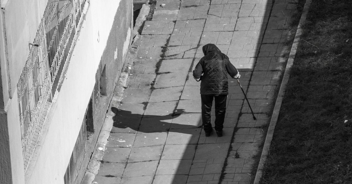 Monochrome image of an elderly person using a cane on a narrow outdoor path, casting a shadow.