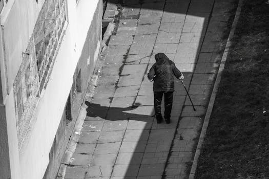Monochrome image of an elderly person using a cane on a narrow outdoor path, casting a shadow.