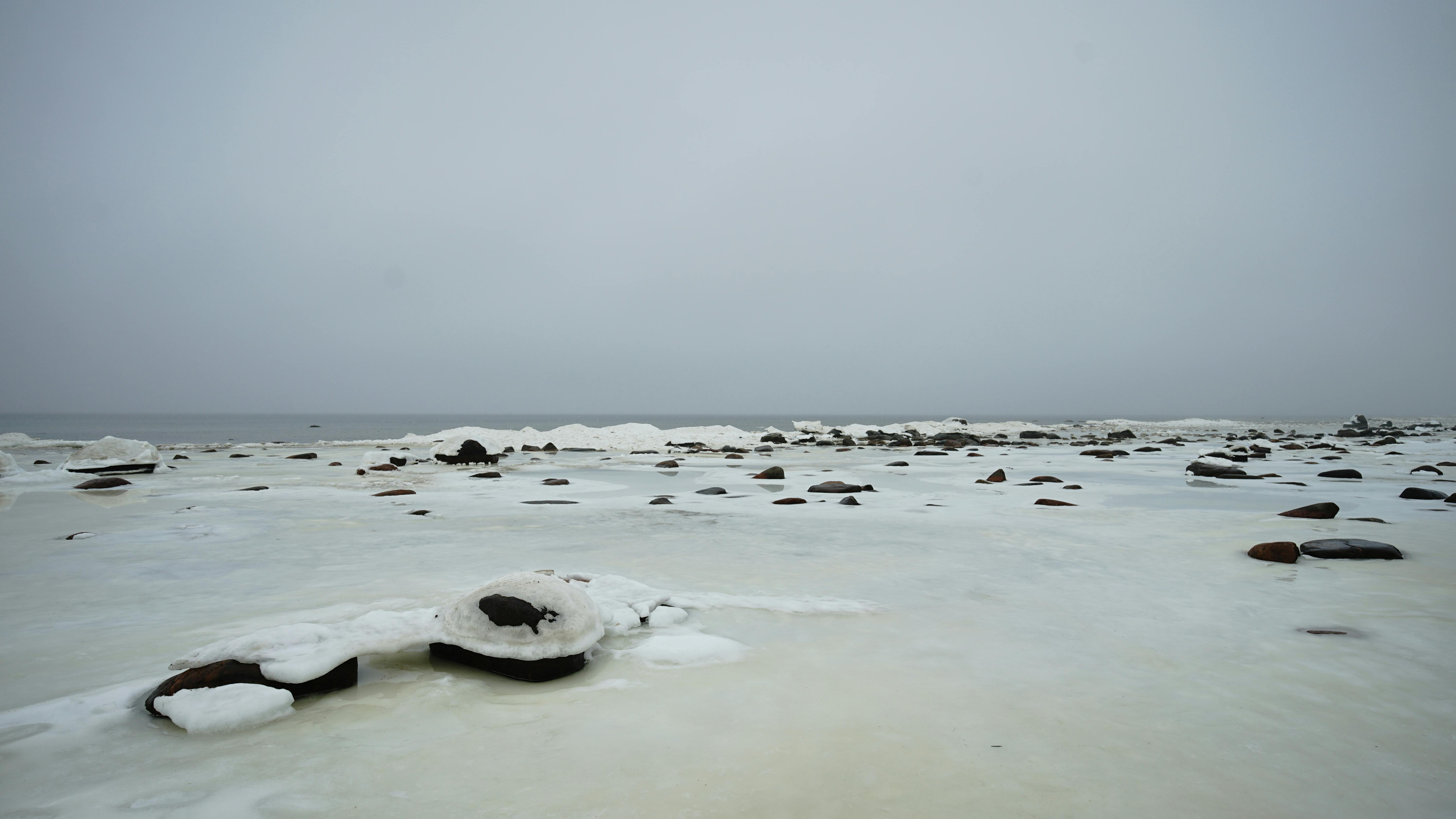 無料 曇り空の下、岩や氷が点在する凍った海岸線の静かな冬の海の景色。 写真素材