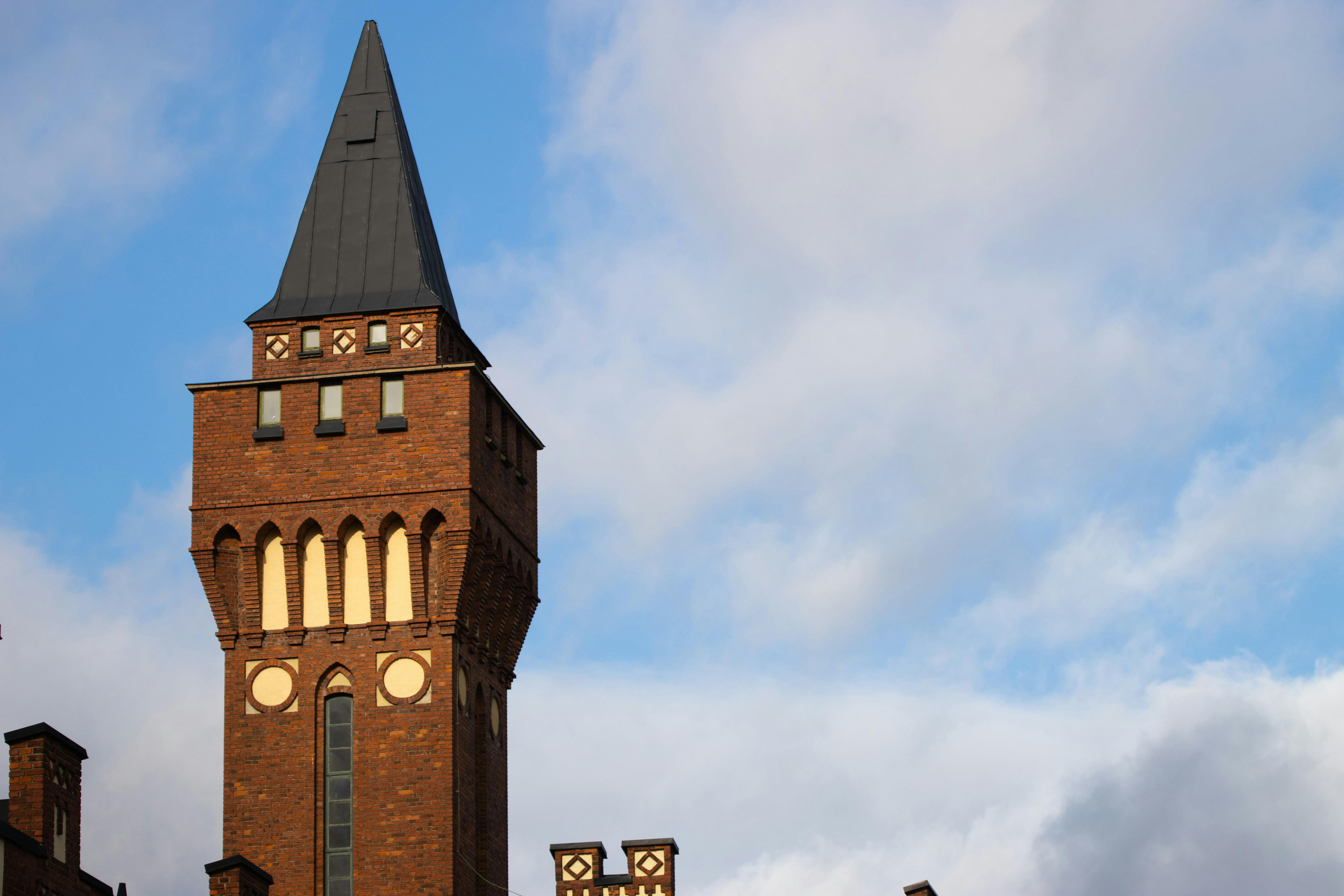 無料 尖った屋根を持つ歴史的なレンガ造りの塔が、点在する雲のある鮮やかな青空を背景に高くそびえ立っています。 写真素材