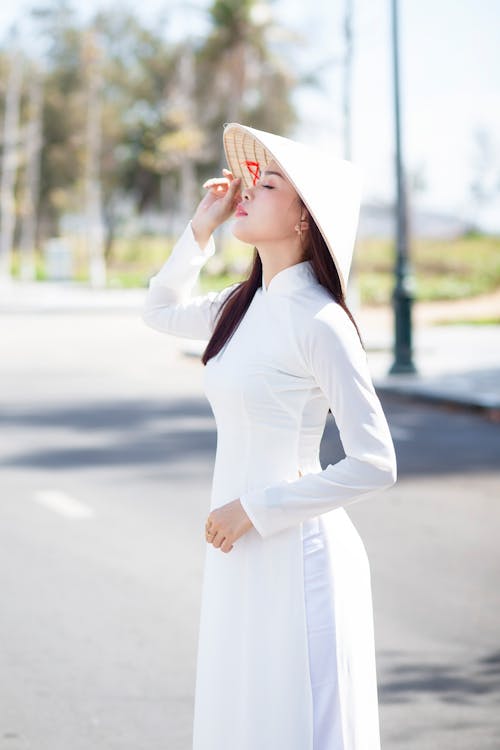 白色長袖連衣裙和白色太陽帽站在路上的女人