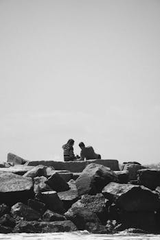 Silhouette of two people seated on coastal rocks against a clear sky.