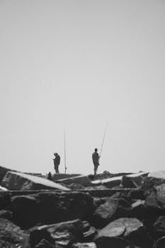 Black and white silhouette of two fishermen standing on a rocky shoreline with fishing rods.