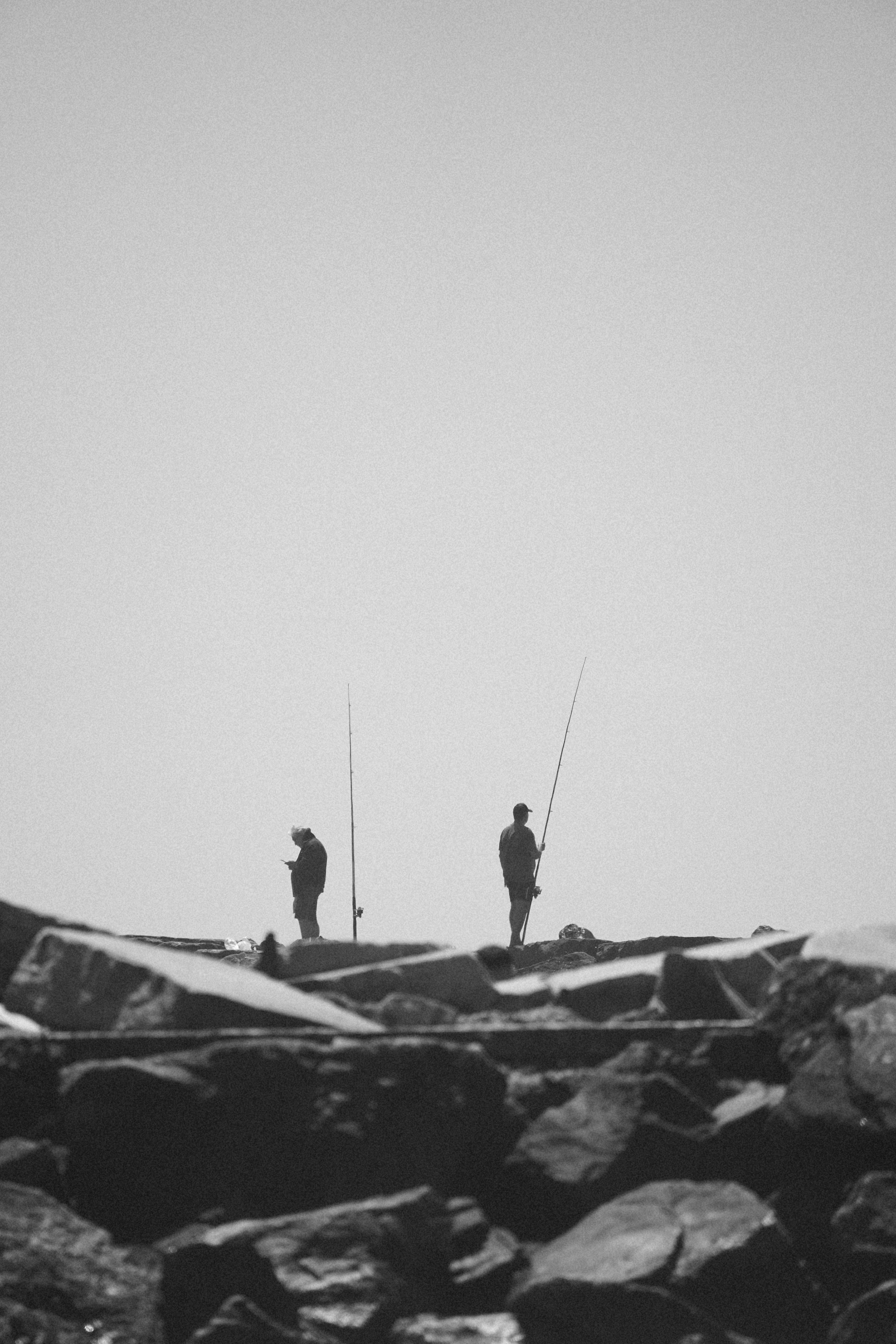 Gratis Silueta en blanco y negro de dos pescadores de pie en una costa rocosa con cañas de pescar. Foto de stock