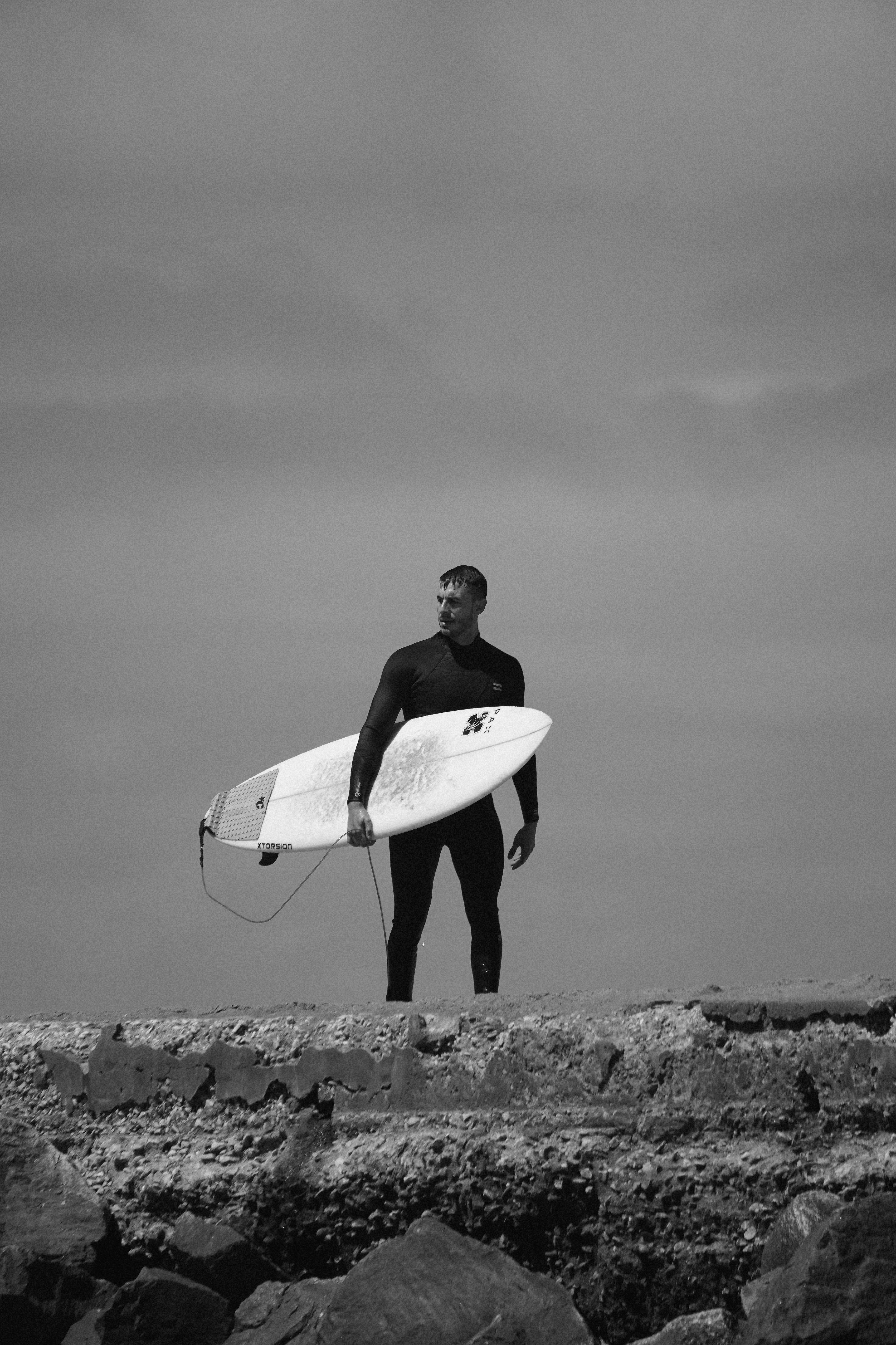 Free A surfer in a wetsuit holds a surfboard on a rocky shore under a cloudy sky, in black and white. Stock Photo