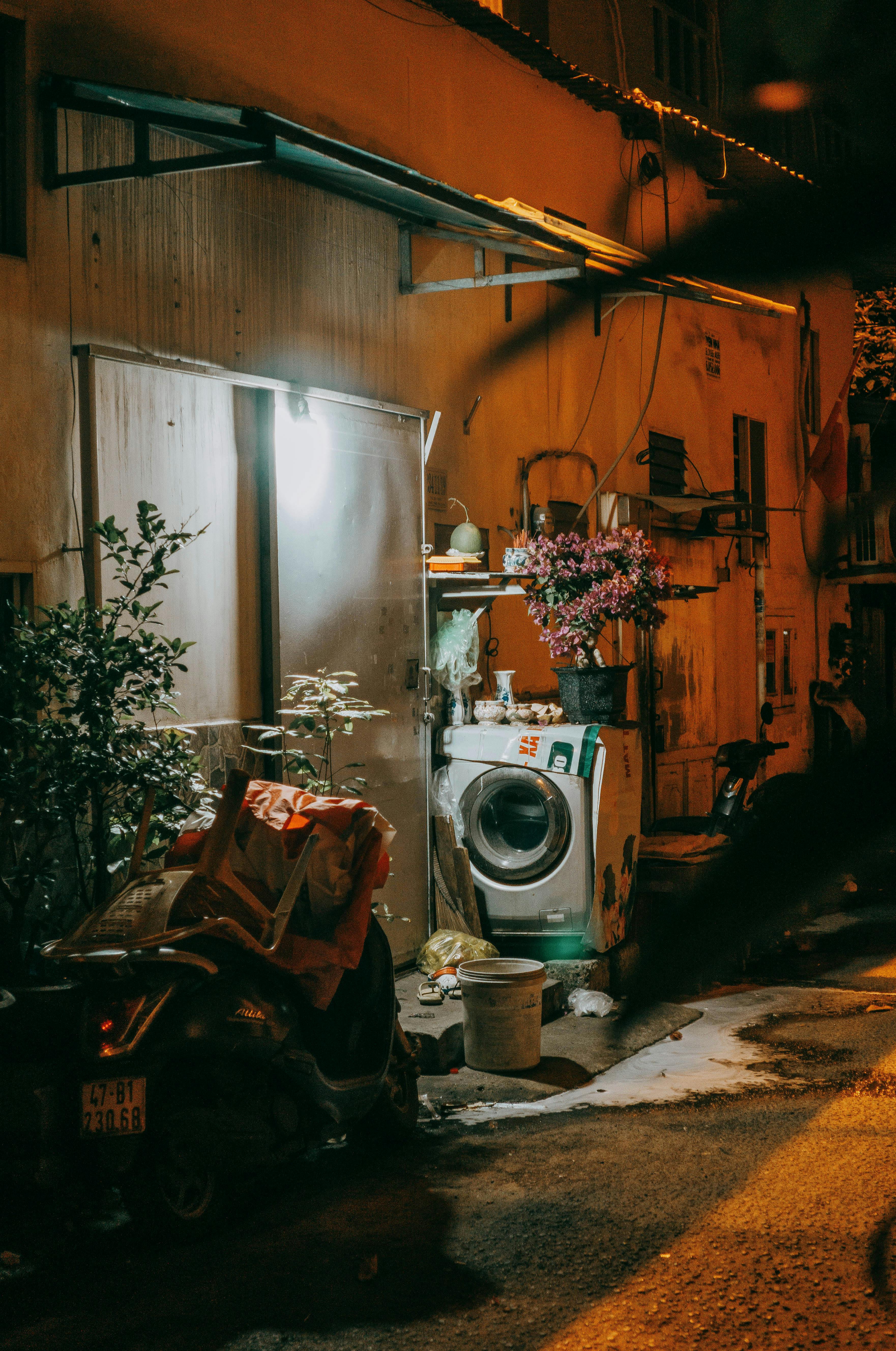無料 洗濯機、植物、スクーターが置かれた、陰鬱な夜の都会の路地。素朴な雰囲気を醸し出しています。 写真素材