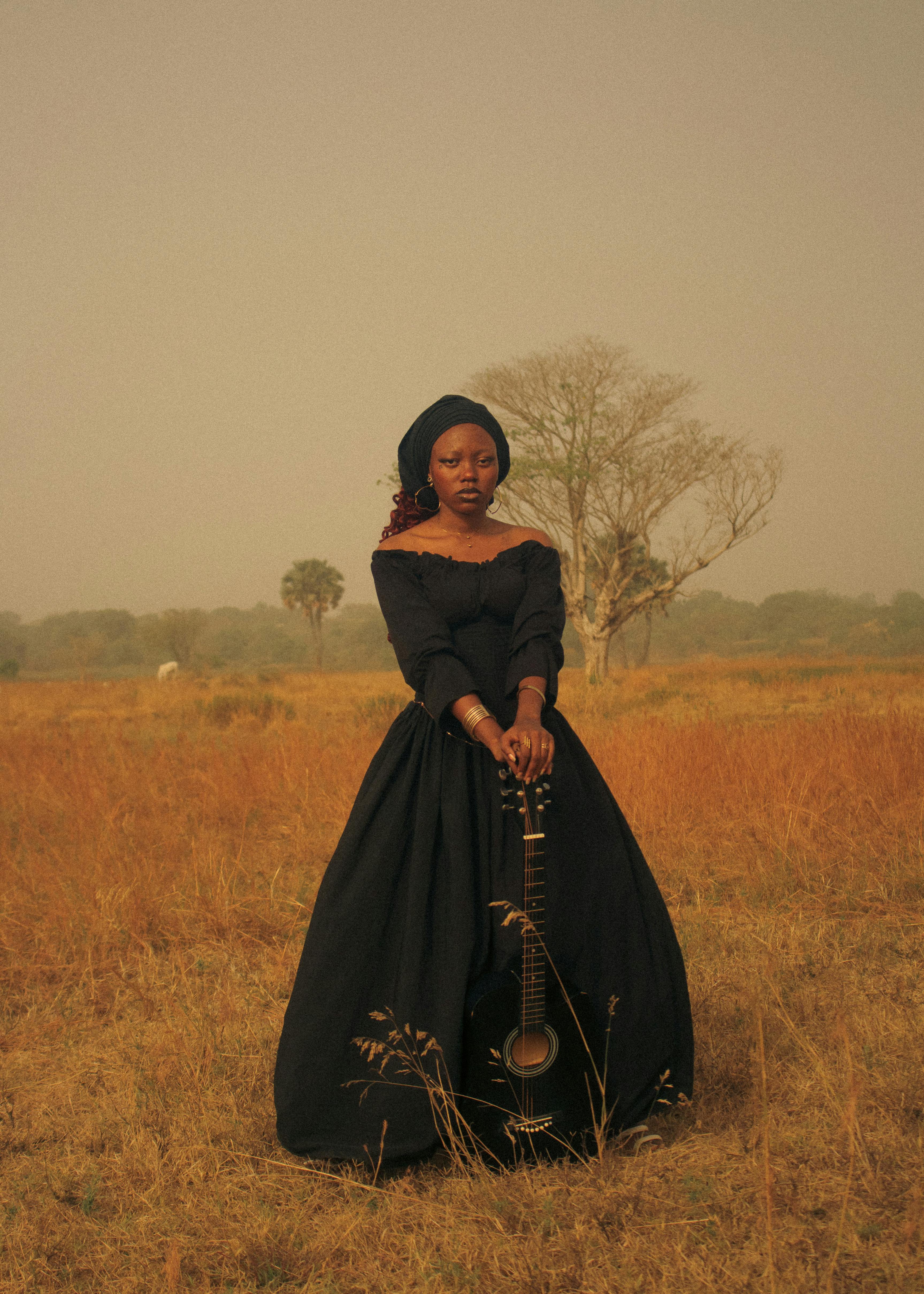 A woman in a black dress stands in a field holding a mandolin, evoking a vintage, earthy mood.