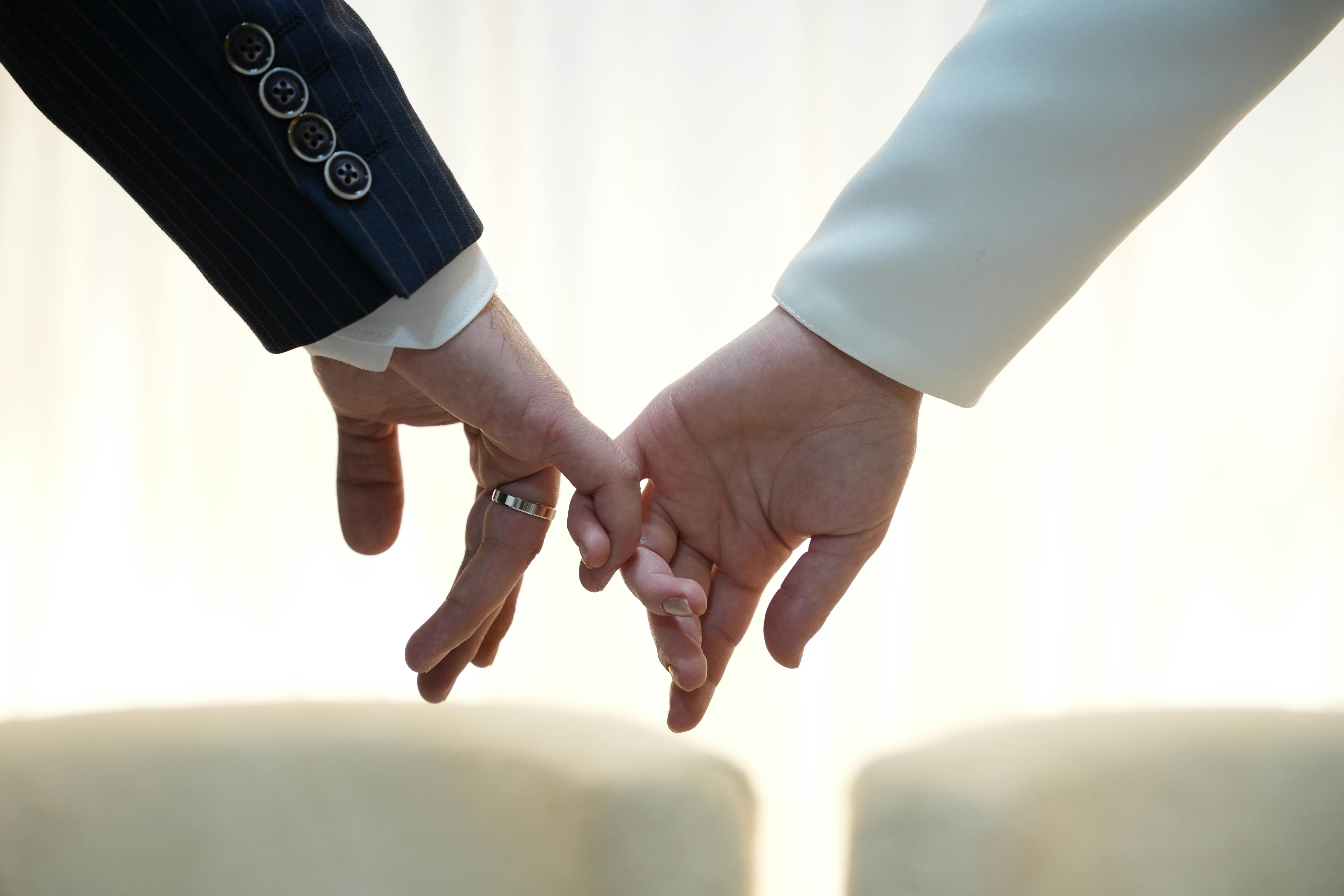 Free A close-up of two adults tenderly holding hands in formal suits, symbolizing unity and connection. Stock Photo
