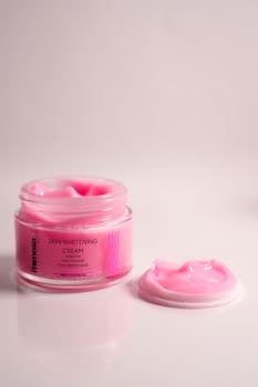 Open jar of pink skin whitening cream with lid on white background.