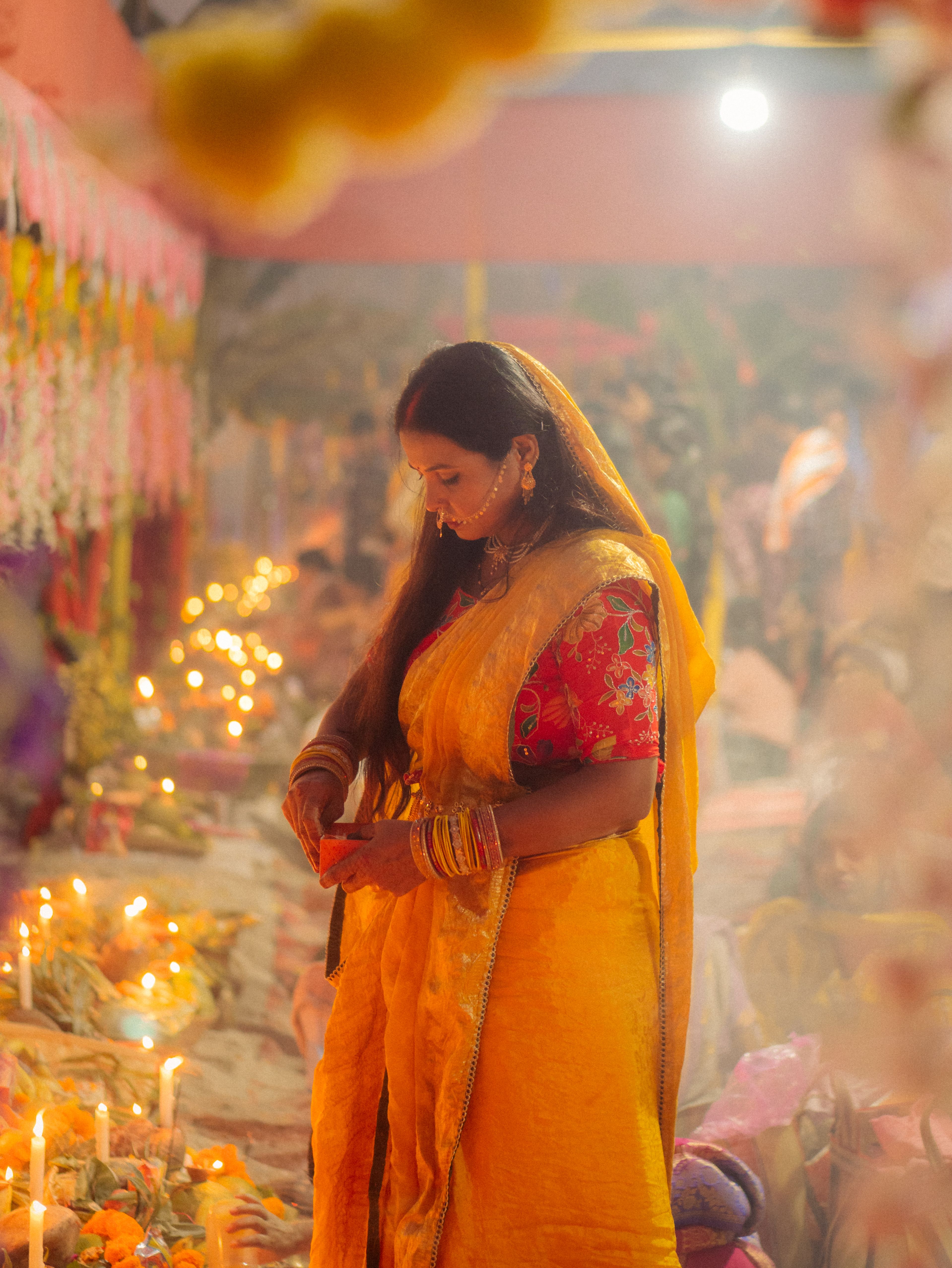 A woman in yellow sari participates in an Indian festive ceremony with candles and flowers.