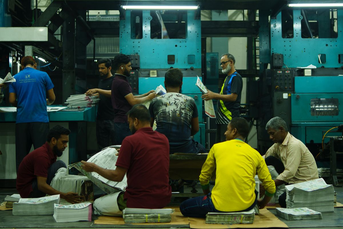Workers sorting bundles of printed newspaper in parallel while the printing presses keep running behind them, one worker left at the machine