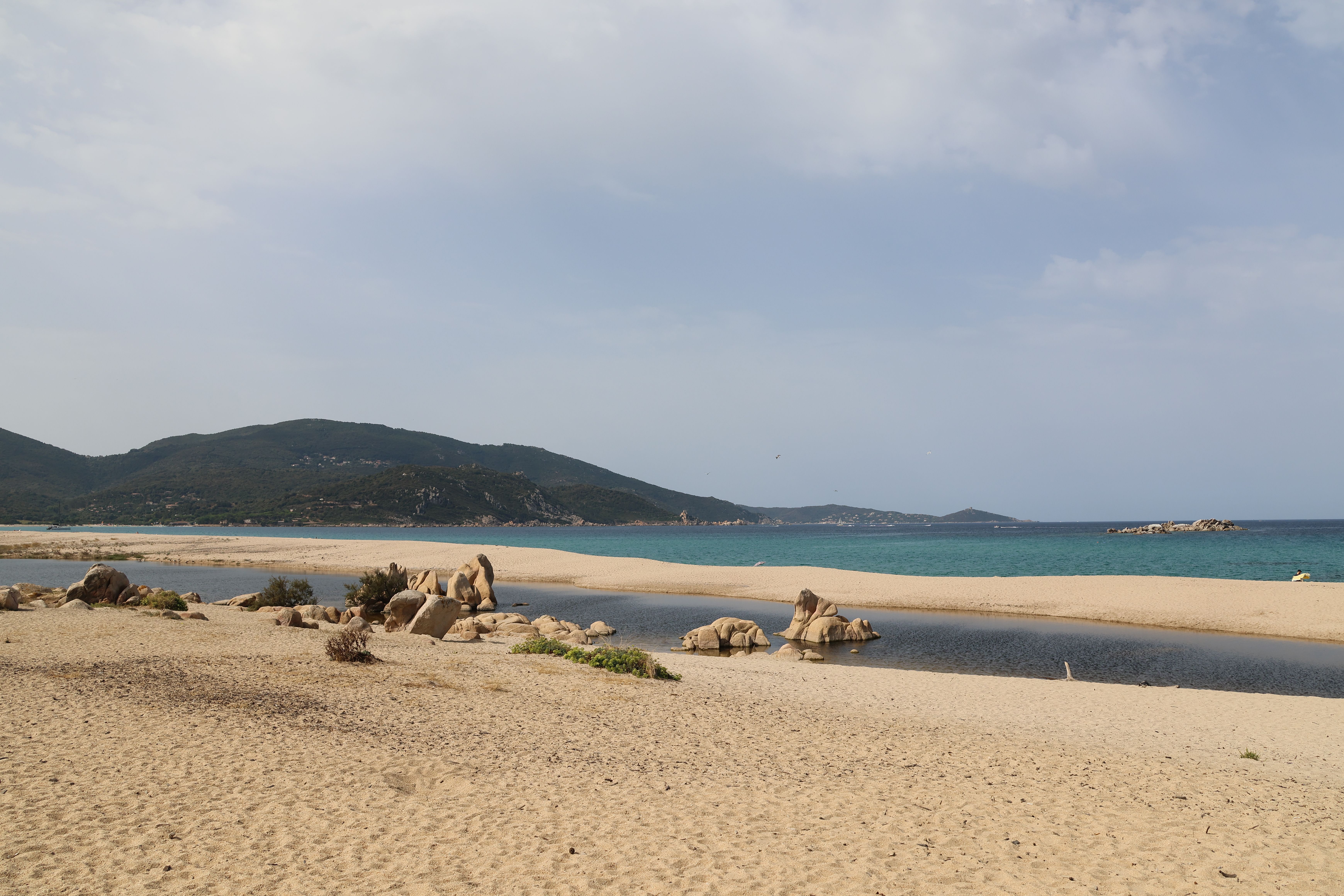 Plage de sable tranquille, rivage rocheux et montagnes au loin sous un ciel nuageux.