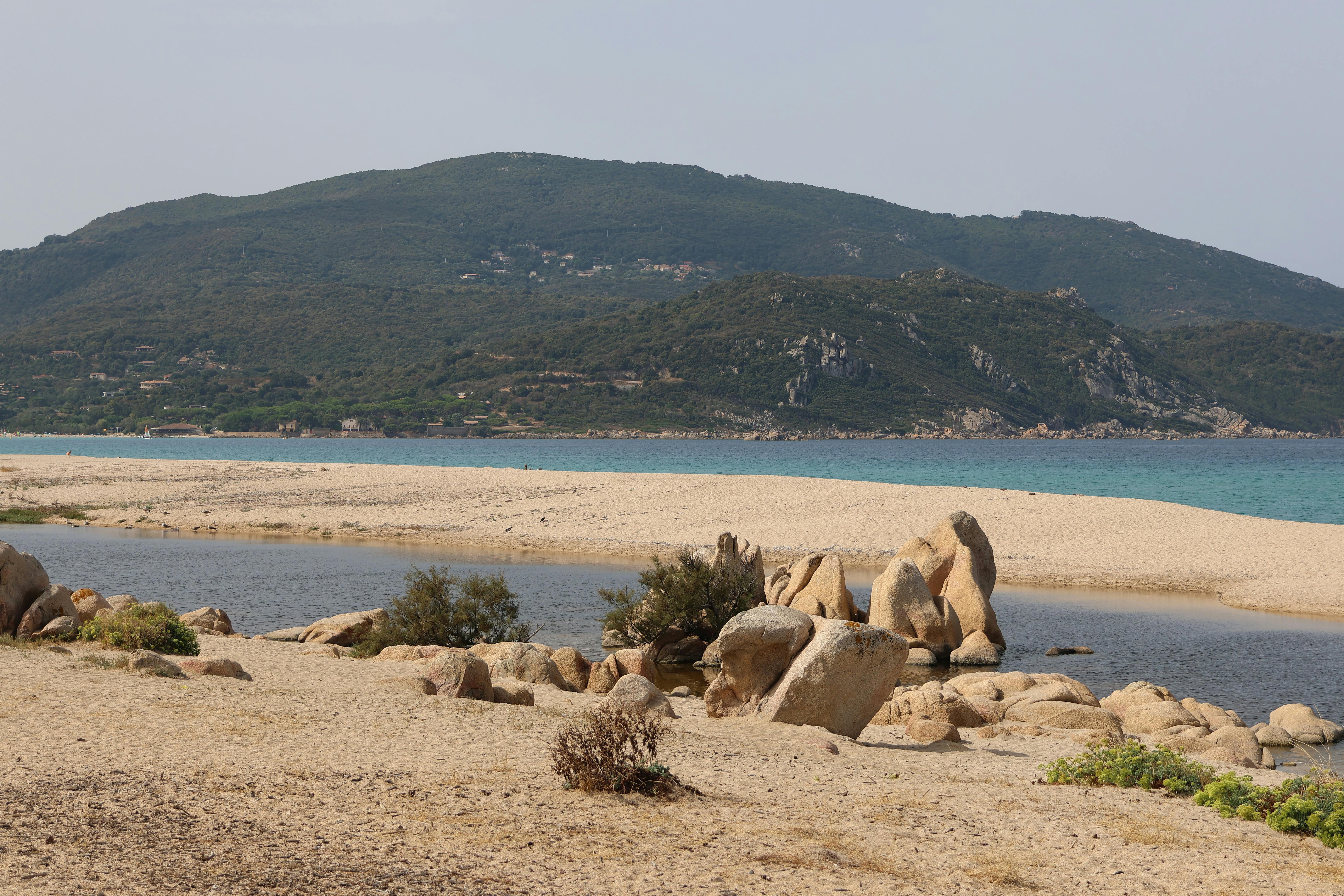Gratis Jelajahi pantai yang tenang dengan formasi batuan dan perbukitan landai di latar belakang. Foto Stok