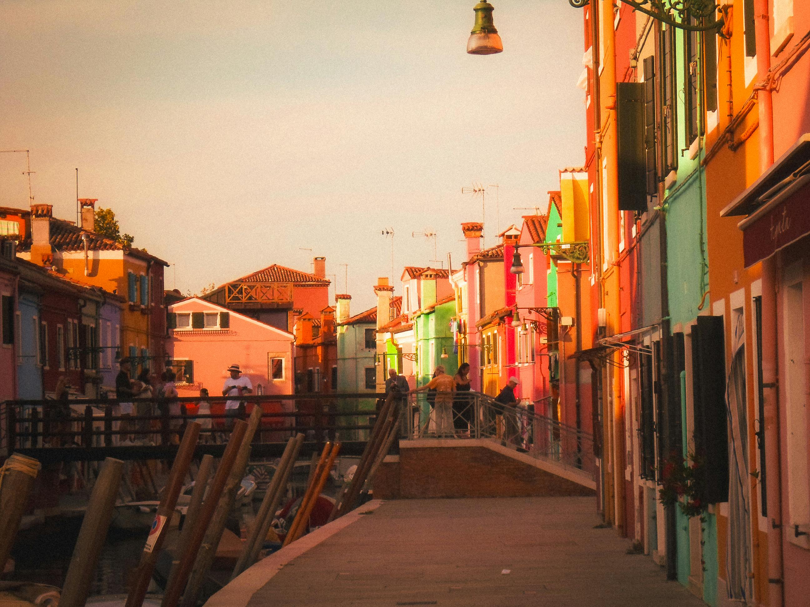 無料 活気のある家々が並び、夕日を眺めながら散歩を楽しむ人々がいる、ヴェネツィアのブラーノ島にある魅力的な通り。 写真素材