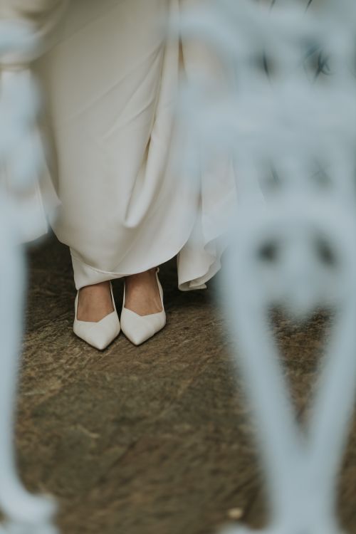 Free Close-up of a bride's elegant white shoes on a rustic wooden floor, framed by metal railing. Stock Photo