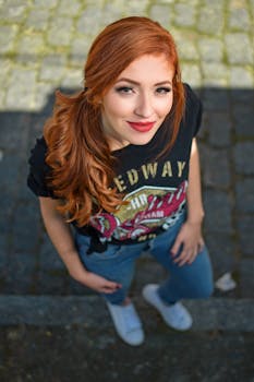A young woman with red hair smiles confidently in a casual outdoor setting.