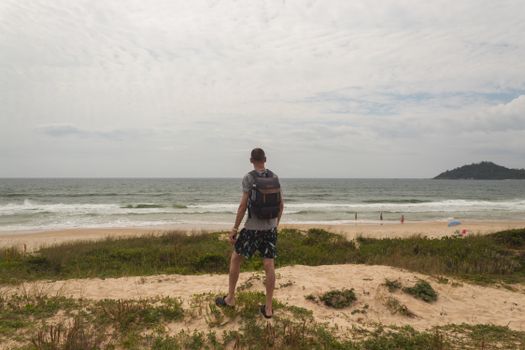 A man with a backpack stands on a hill overlooking a beach, capturing the serene ocean view.