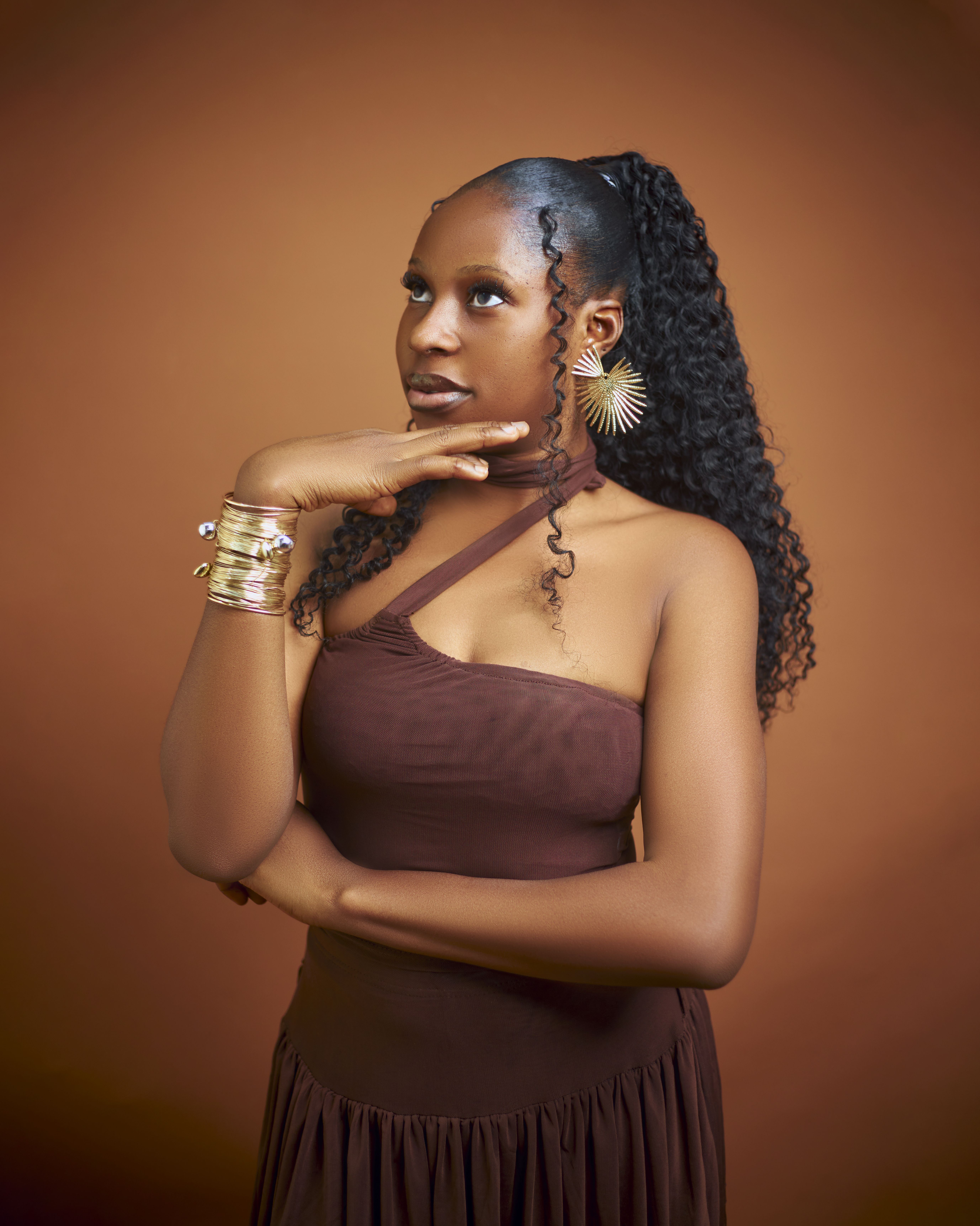 Stylish woman in a brown dress against a warm, brown background looking thoughtful.