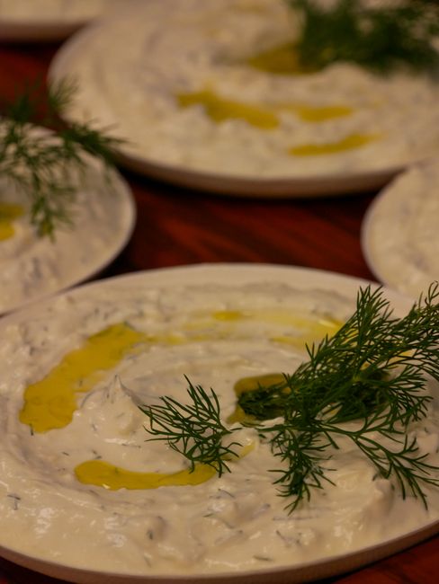 A white bowl of pale green tzatziki dip swirled with olive oil and flecked with fresh dill, surrounded by wedges of warm pita and cucumber slices.
