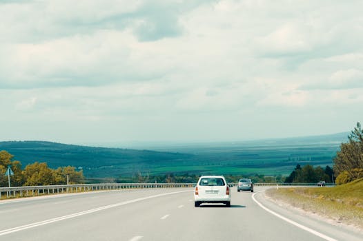 A serene view of a road with cars, bordered by lush landscapes under a cloudy sky.