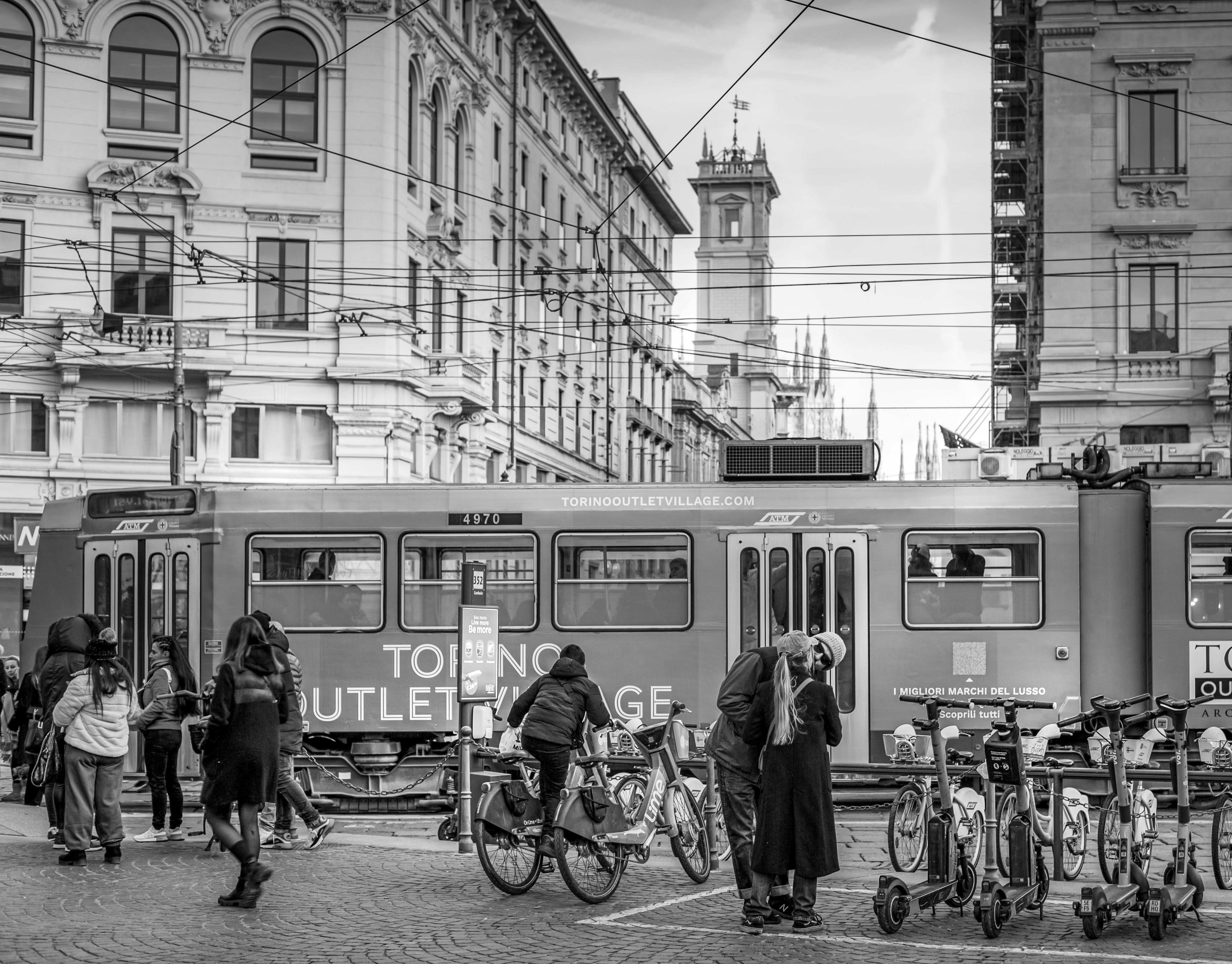 A lively street scene in Milano, showing people with bicycles and a tram in a bustling urban environment.