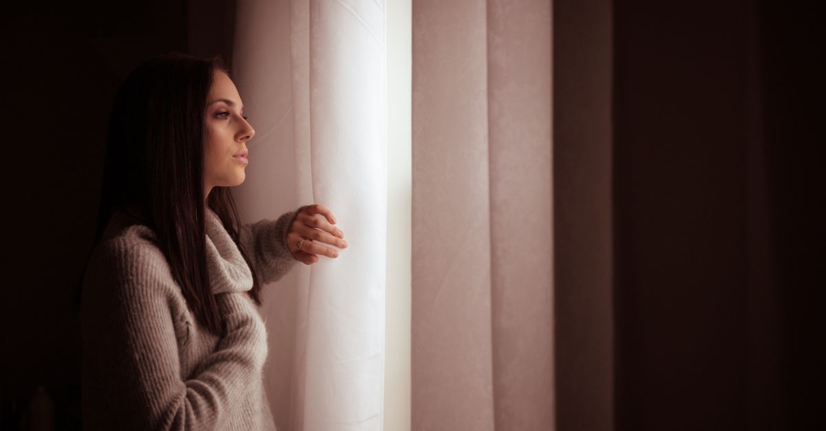 A woman in a sweater thoughtfully gazes out of a sunlit window, creating a warm indoor atmosphere.