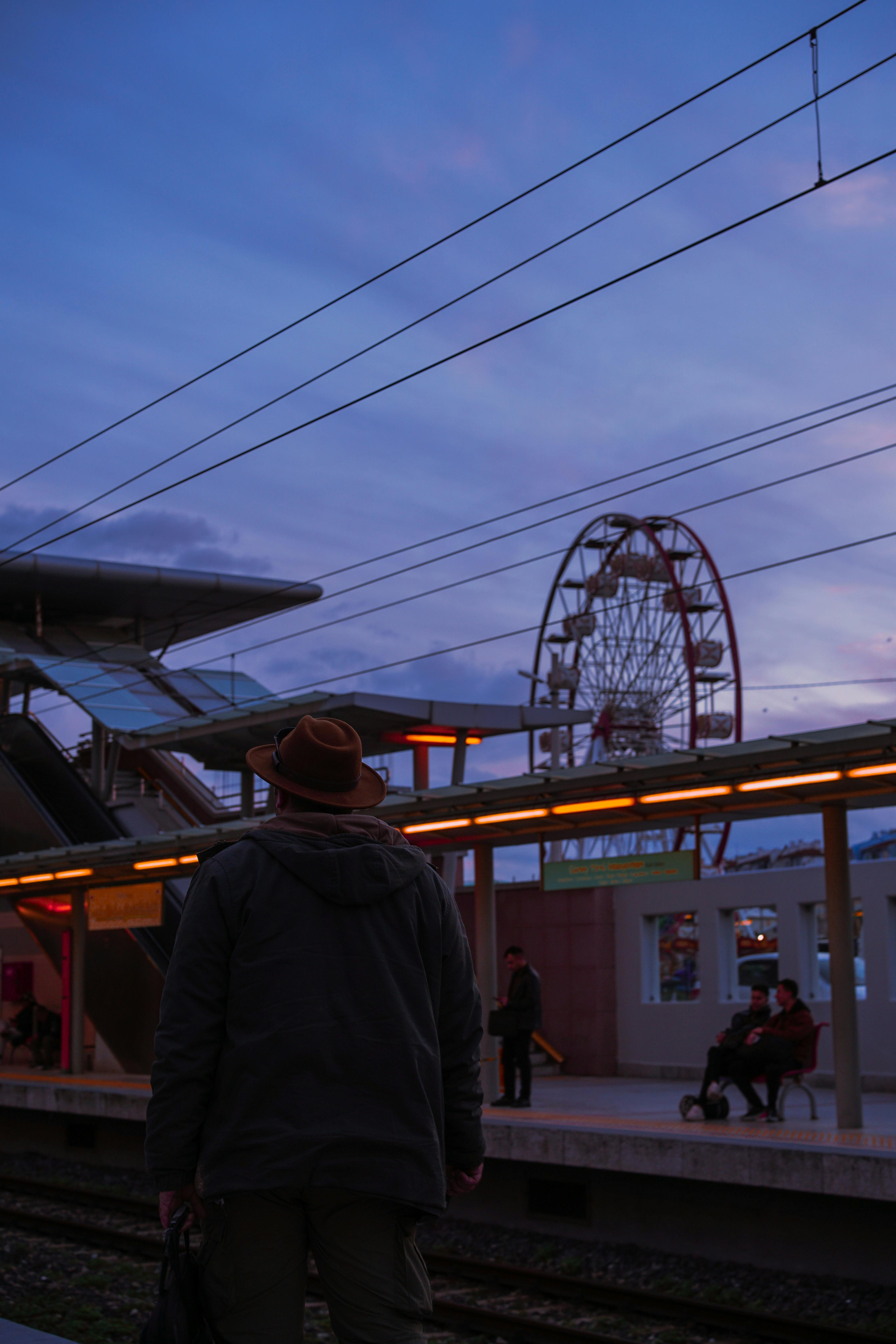 無料 夕暮れ時、帽子をかぶった男性が駅のプラットフォームで点灯した観覧車を眺めている。 写真素材