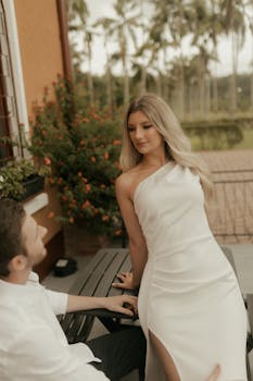 A couple seated on a bench outdoors, dressed elegantly amidst lush greenery.