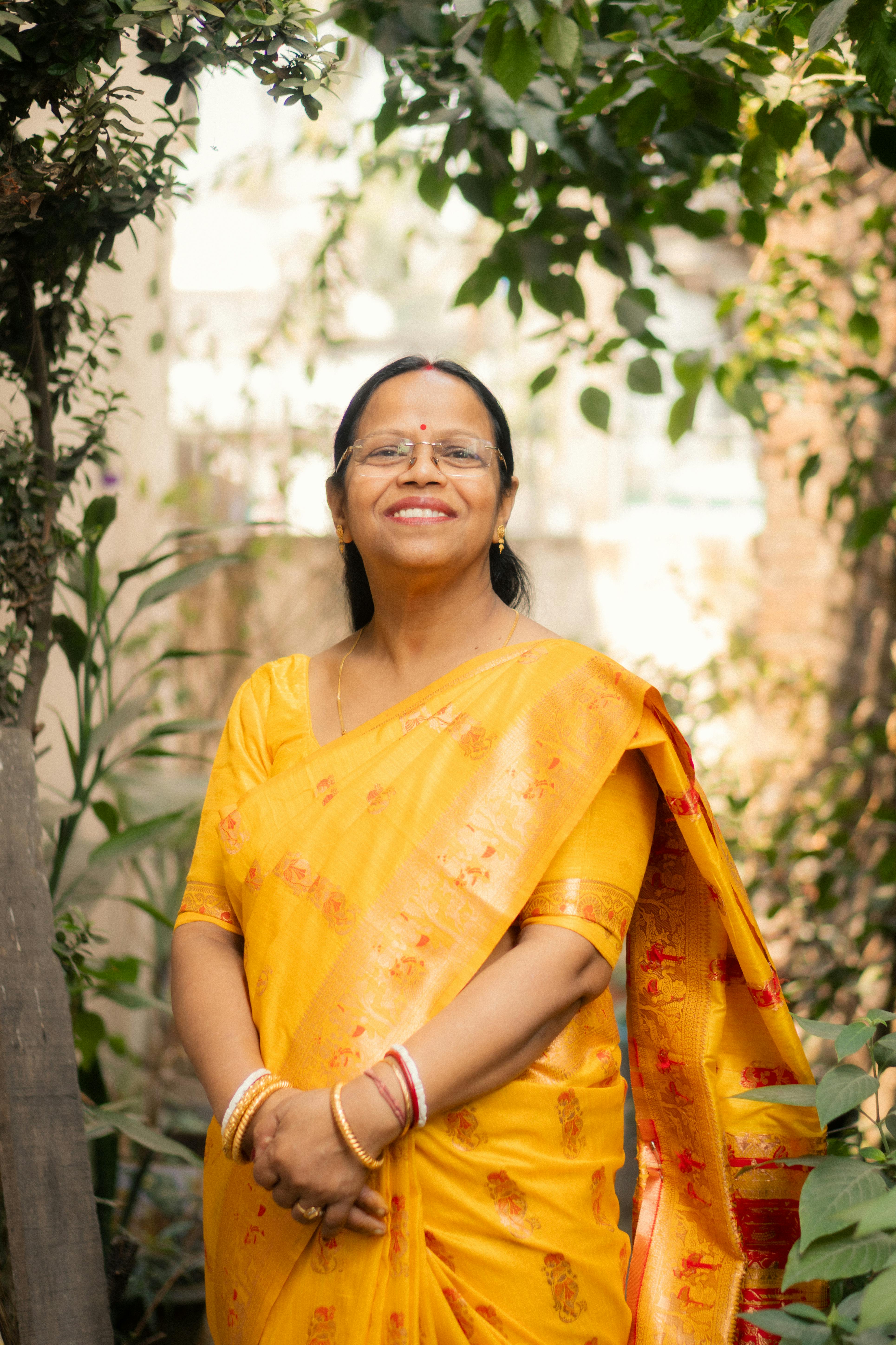 Portrait of a smiling woman in a yellow saree, standing outdoors with greenery around.