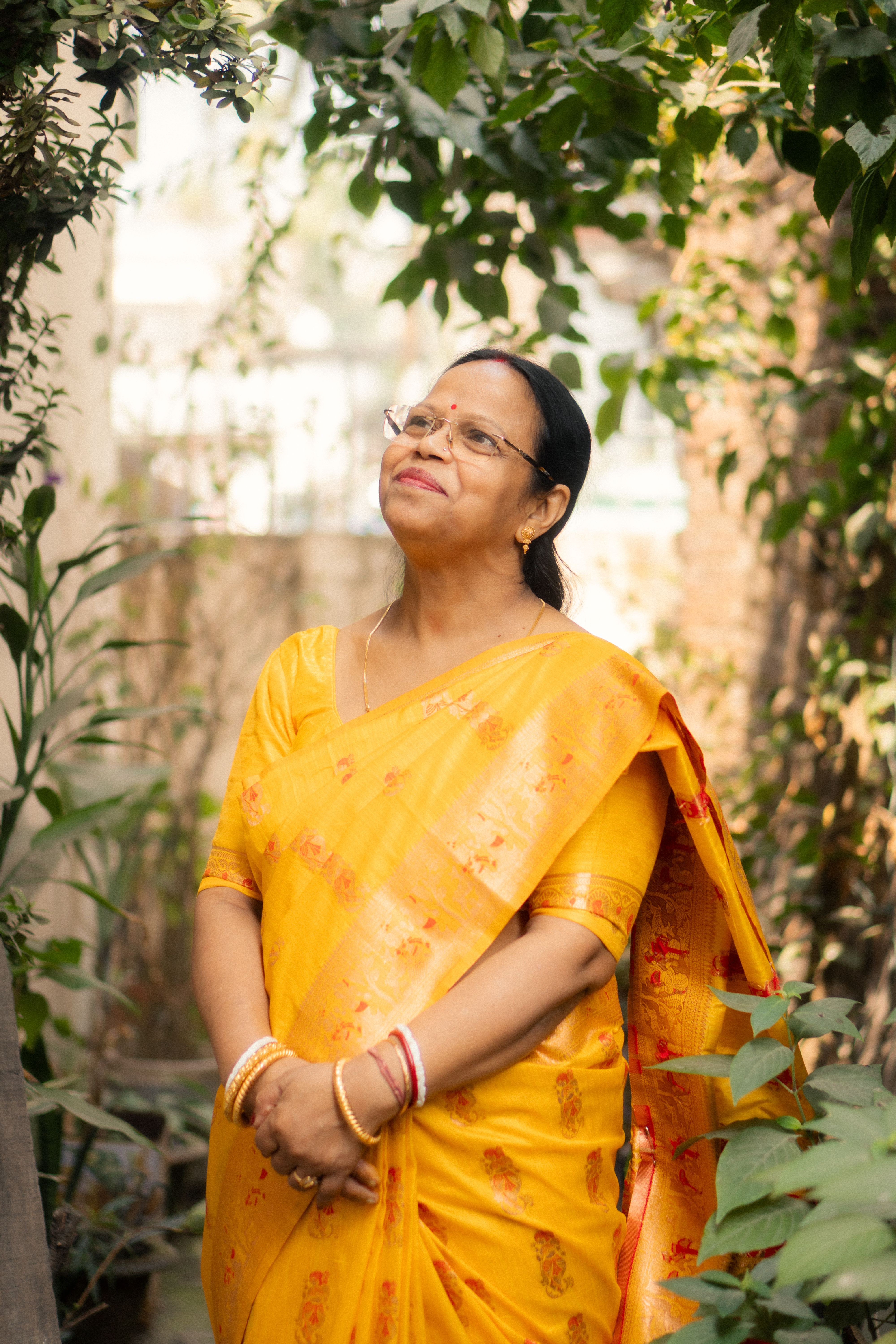 A cheerful woman in a yellow saree enjoys a serene outdoor moment surrounded by greenery.