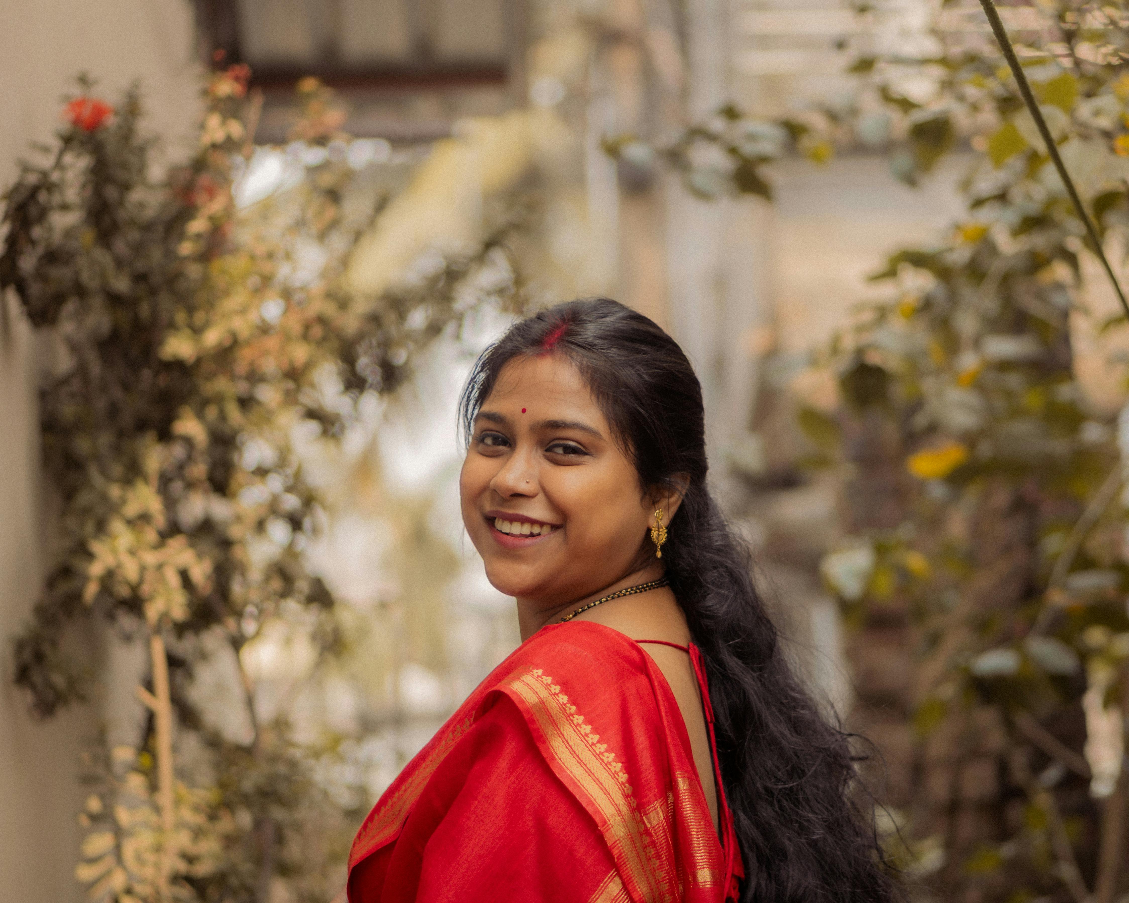 Smiling woman wearing vibrant red saree poses outdoors amidst greenery, showcasing cultural elegance.