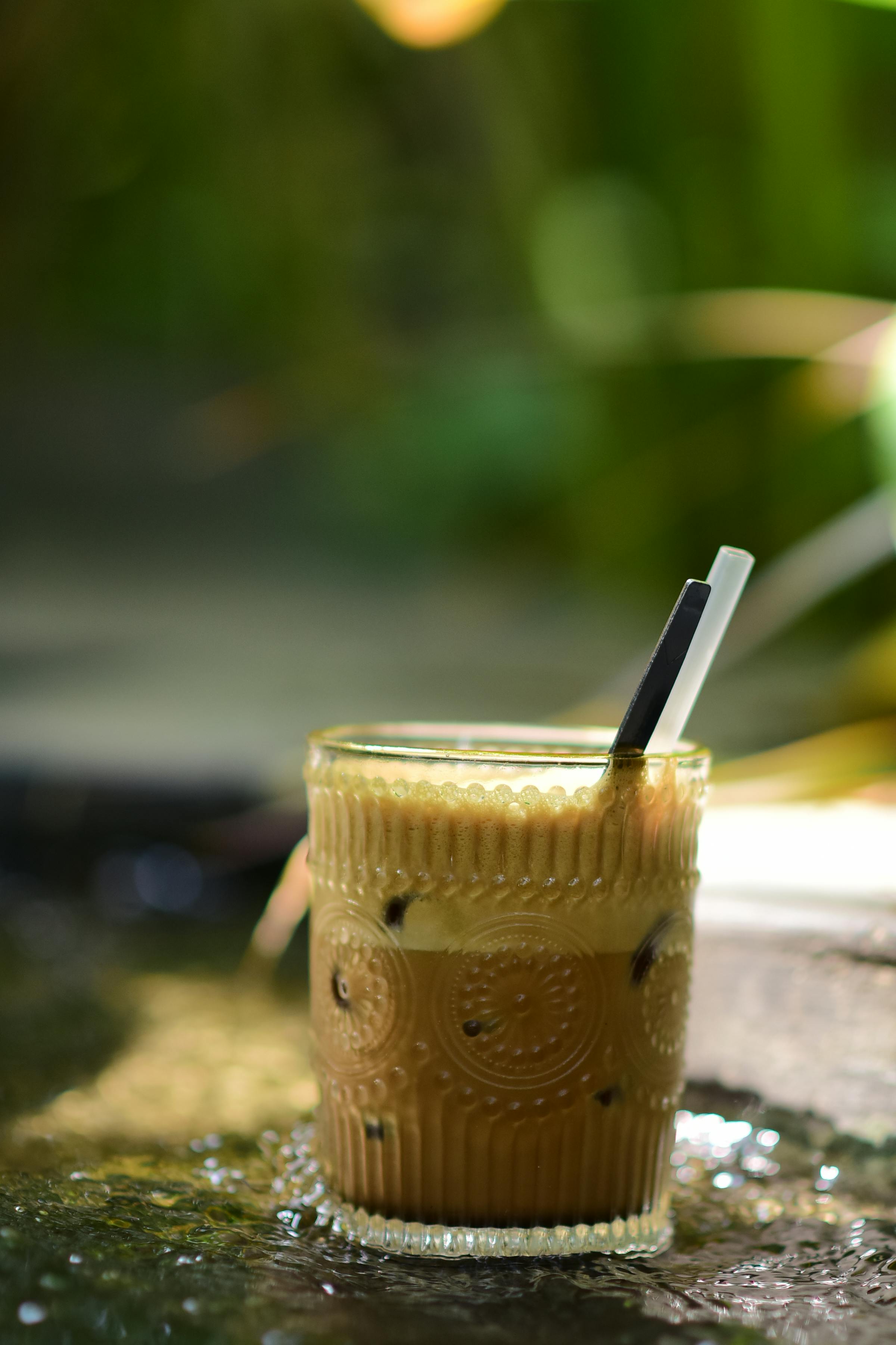 Free A glass of iced coffee with straws sits on wet stones under sunlight, creating a refreshing scene. Stock Photo