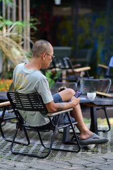 Casual man sitting in an outdoor cafe garden, using a smartphone and enjoying a coffee.