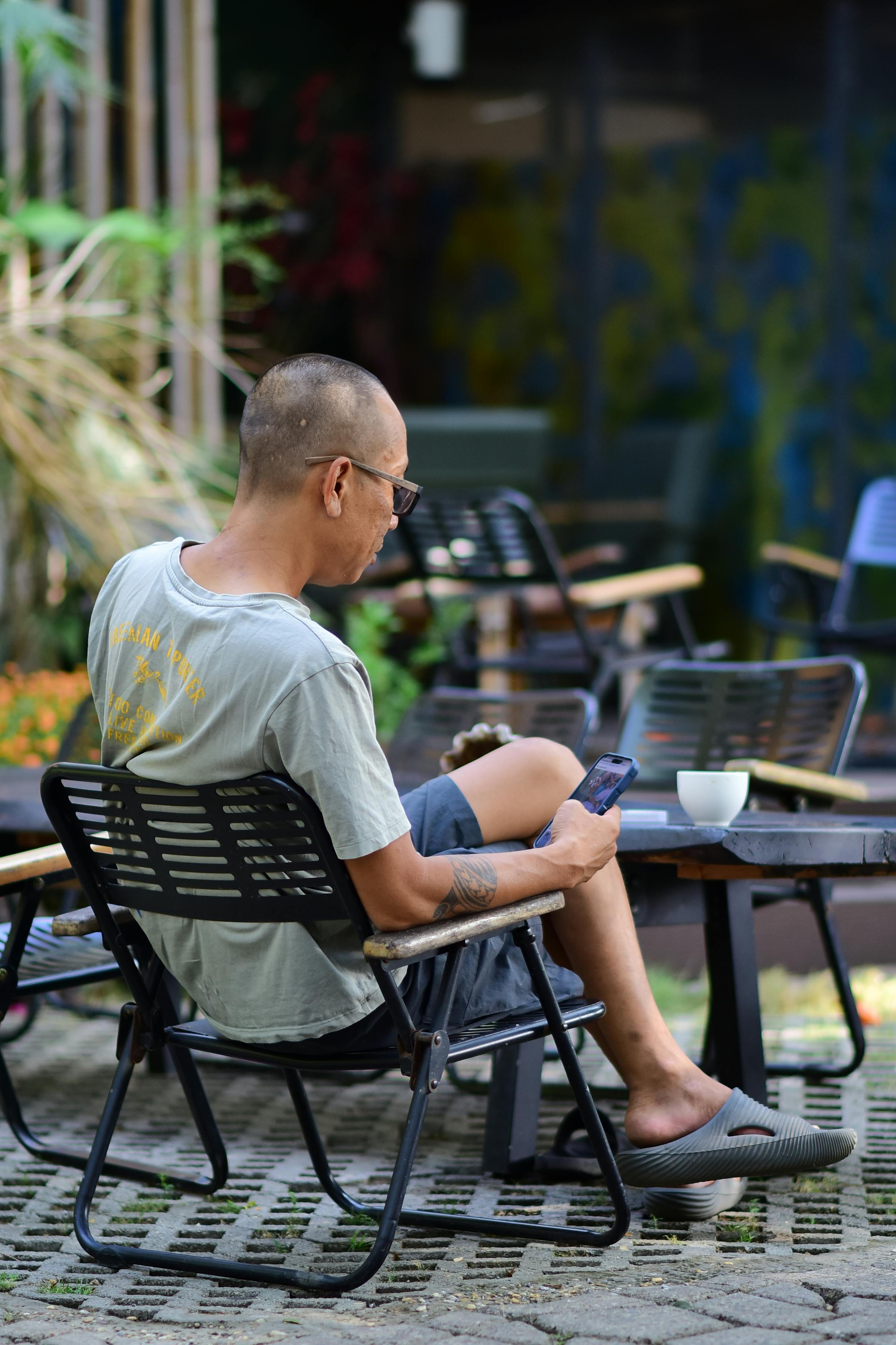 Free A man sits in an outdoor cafe, using his smartphone while enjoying a coffee. Stock Photo