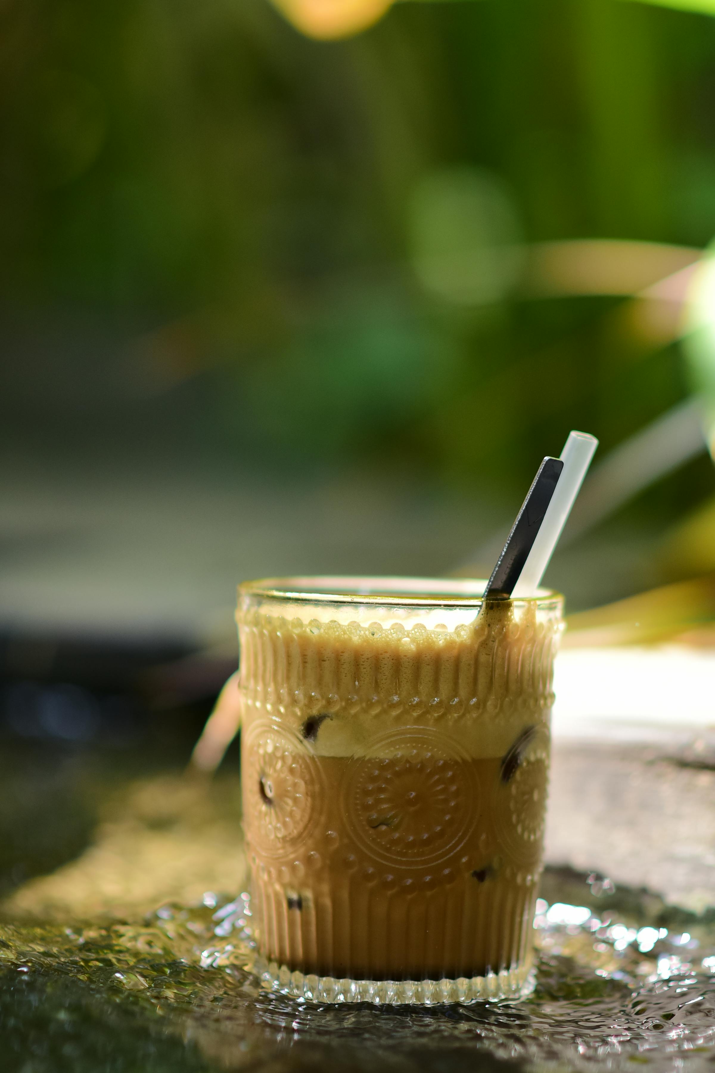 Free A glass of iced coffee with a straw, set against a natural outdoor backdrop, evoking a refreshing and casual vibe. Stock Photo