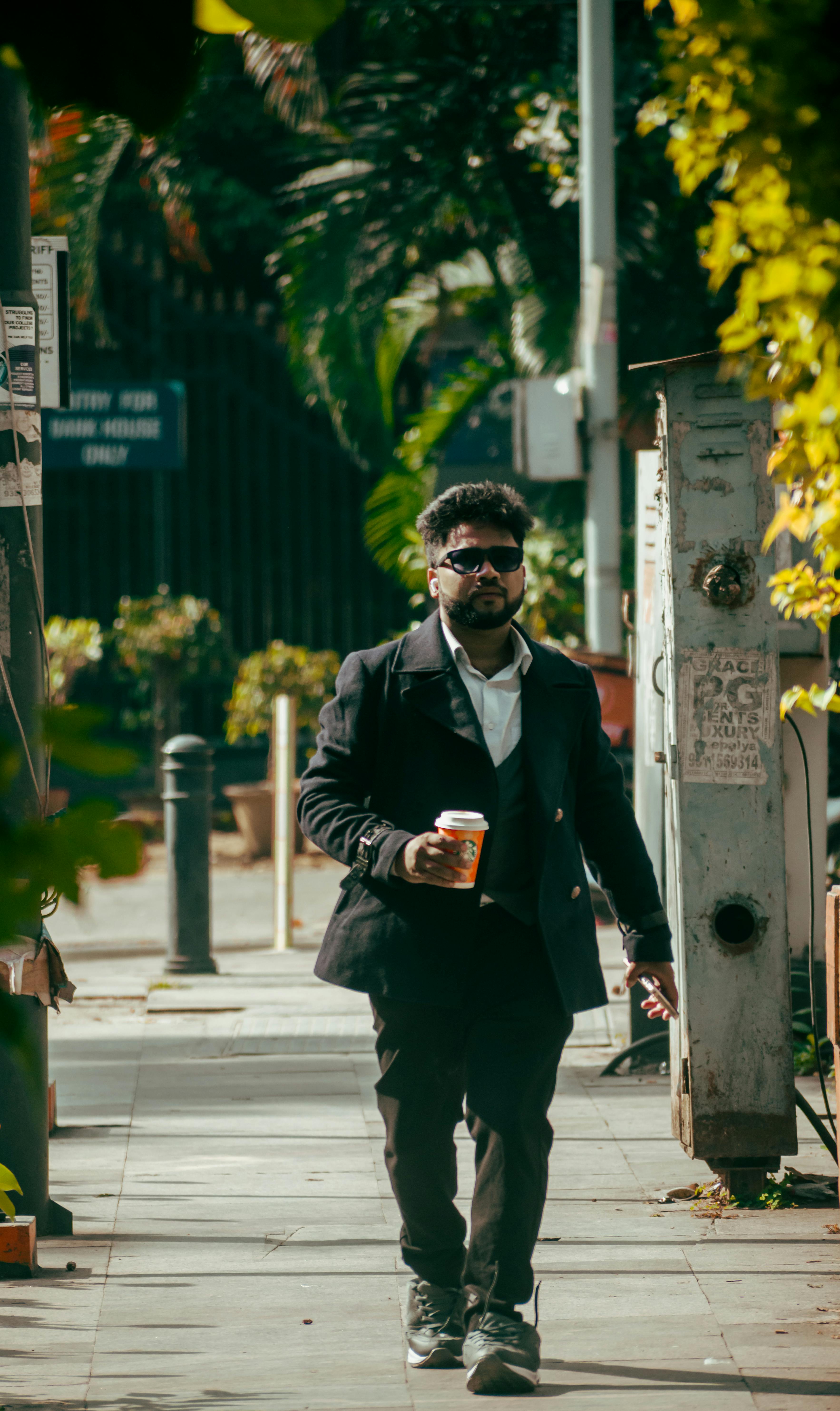 Free A businessman in a suit walking outdoors holding a coffee cup, surrounded by greenery in Bengaluru, India. Stock Photo