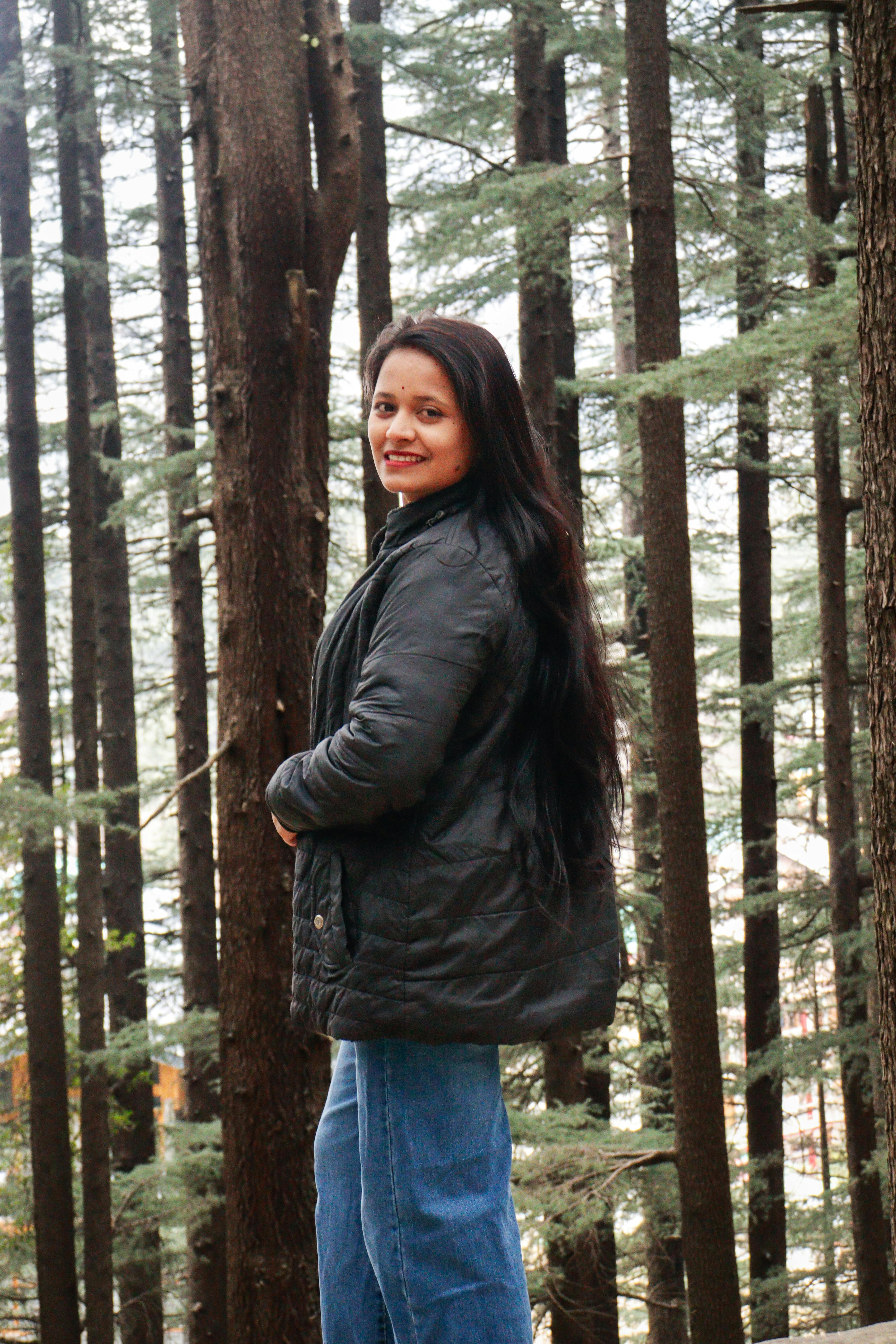 A woman in a jacket smiling in a forest setting with tall trees.