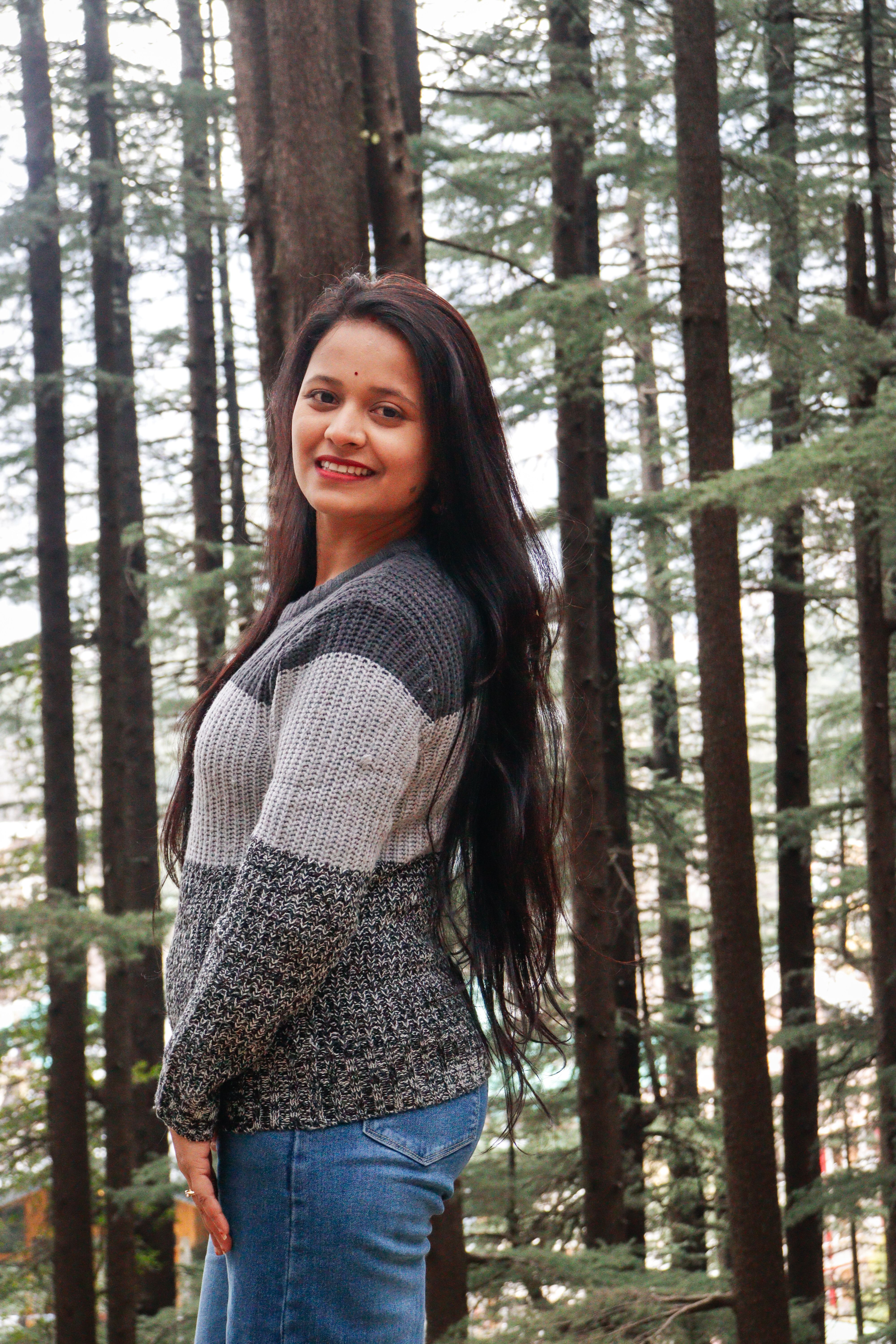A cheerful woman in a cozy sweater standing amidst tall pine trees.