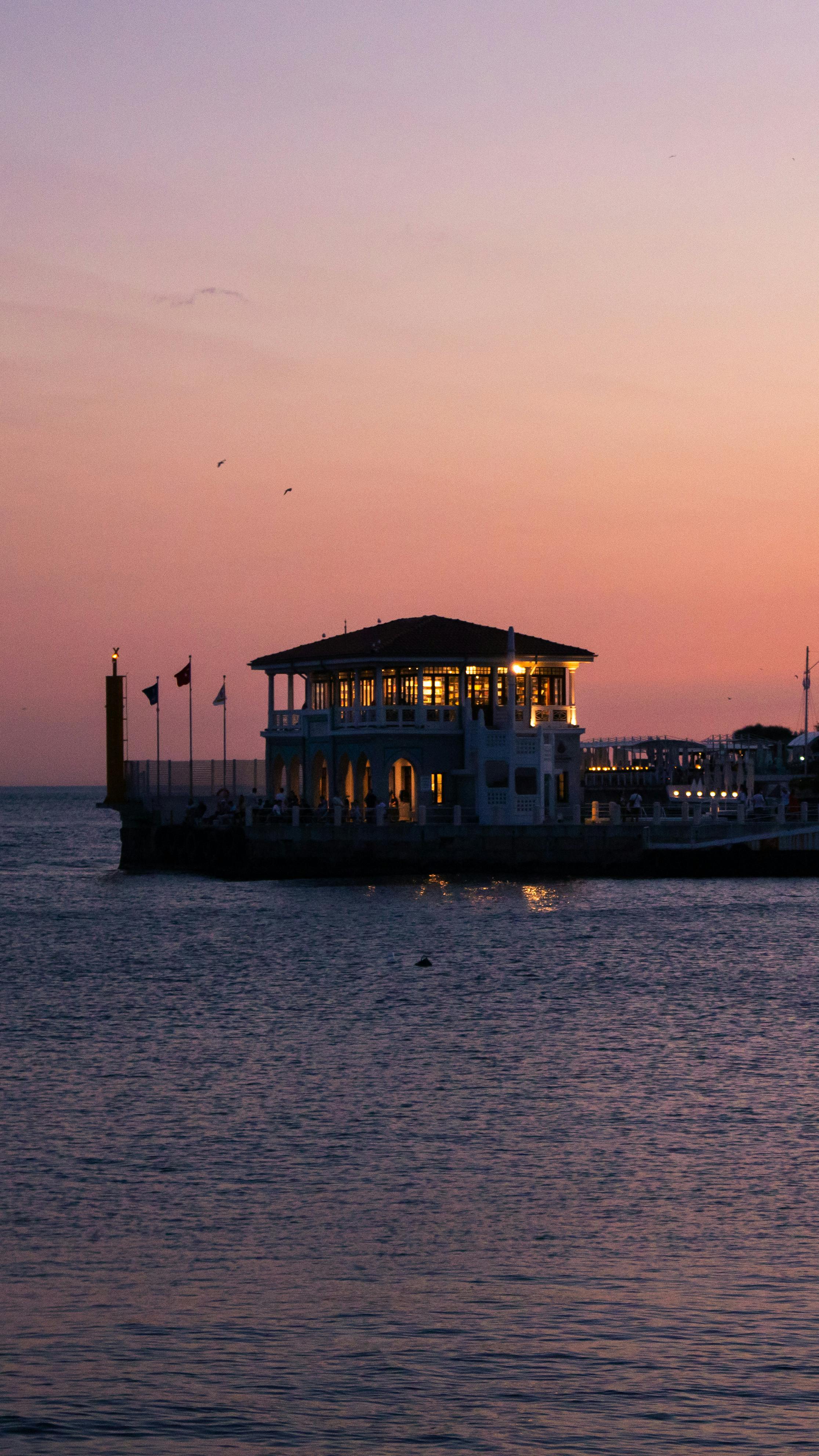 無料 穏やかな夕日と水辺のパビリオンのシルエットが、温かみのある色合いで美しく表現されています。 写真素材