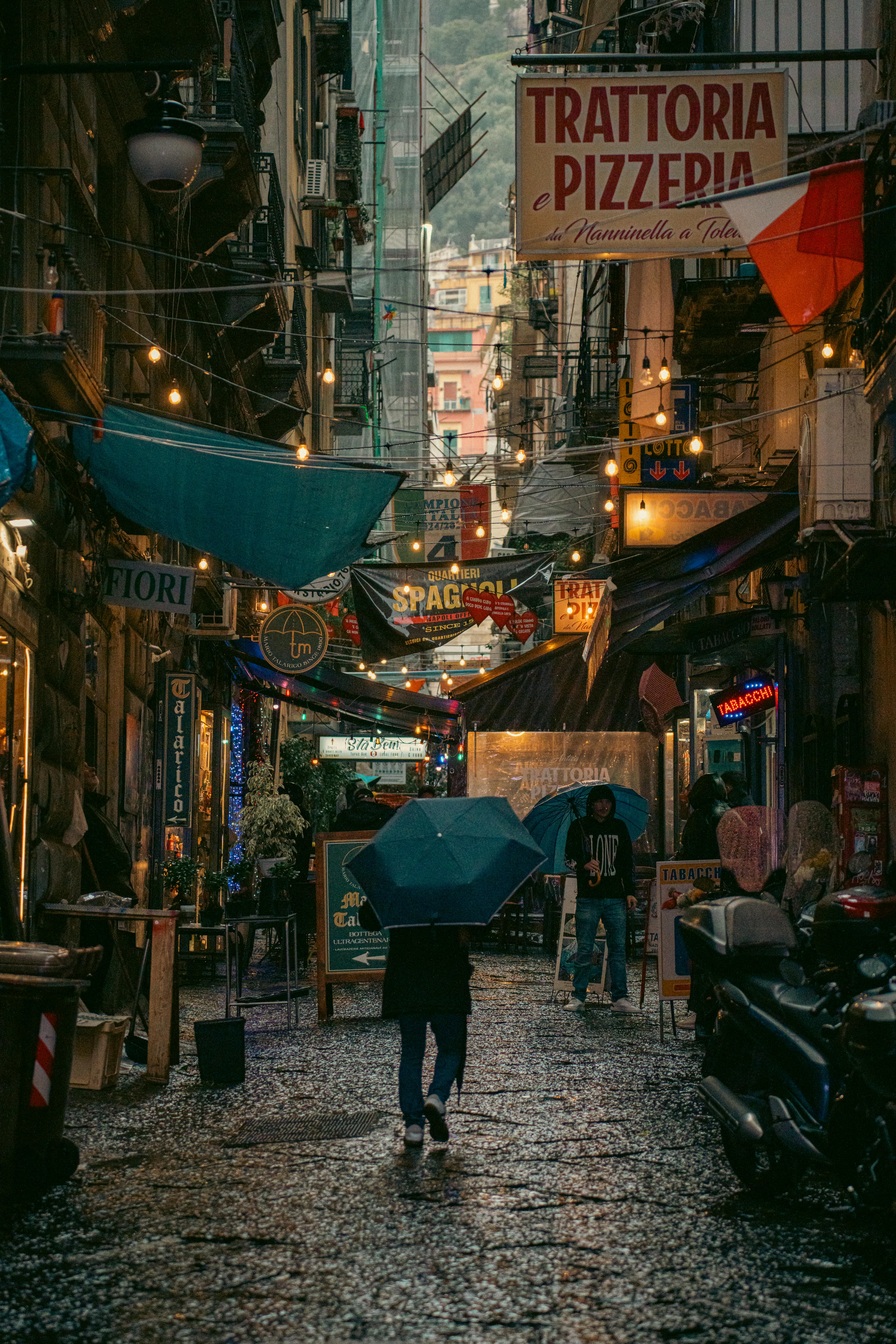 бесплатная Вечерняя прогулка в дождливую погоду по живописному переулку в Неаполе, Италия, с яркими вывесками и зонтиками. Стоковое фото