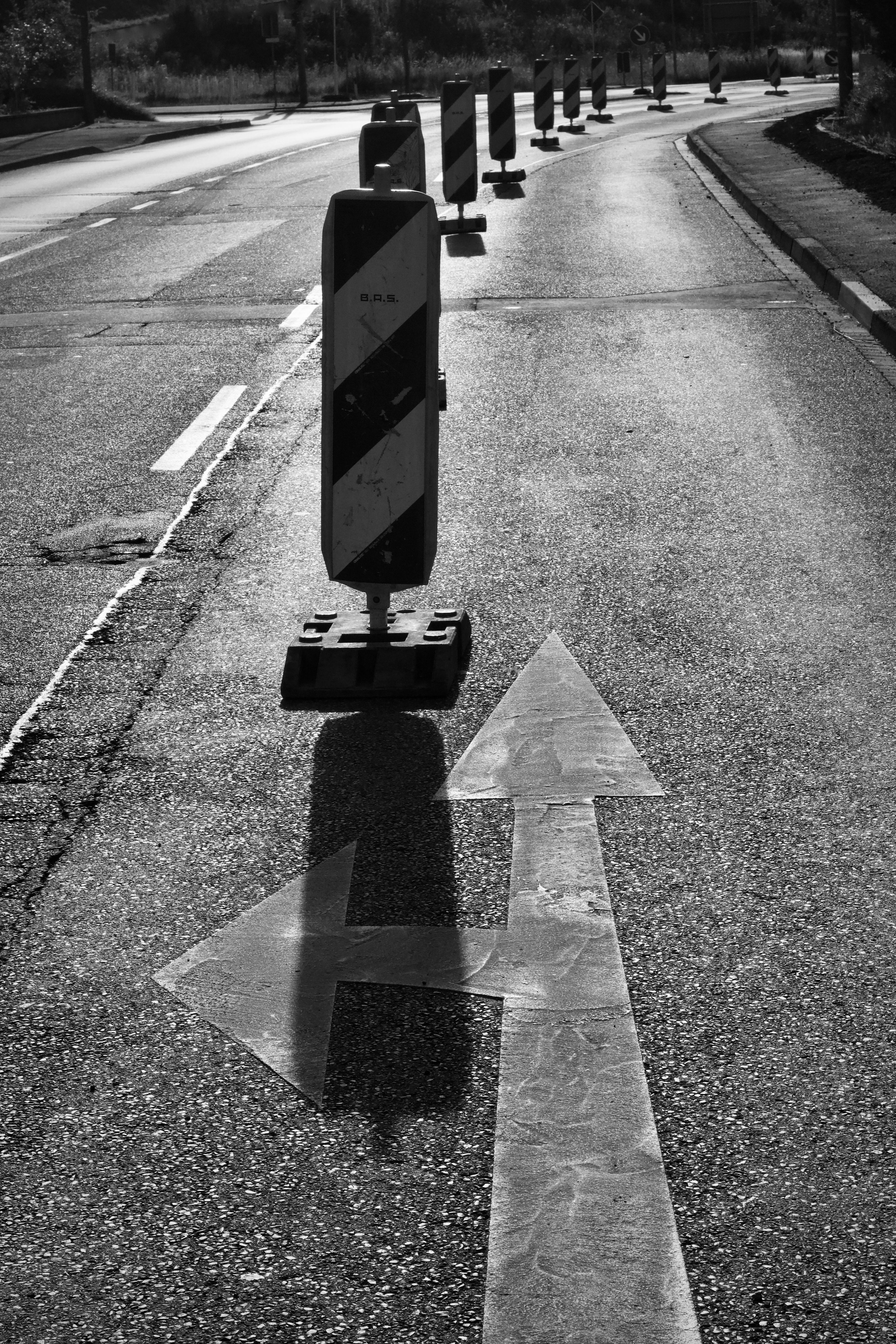 Black and white image of a road with traffic barrier and directional arrows, indicating road construction.