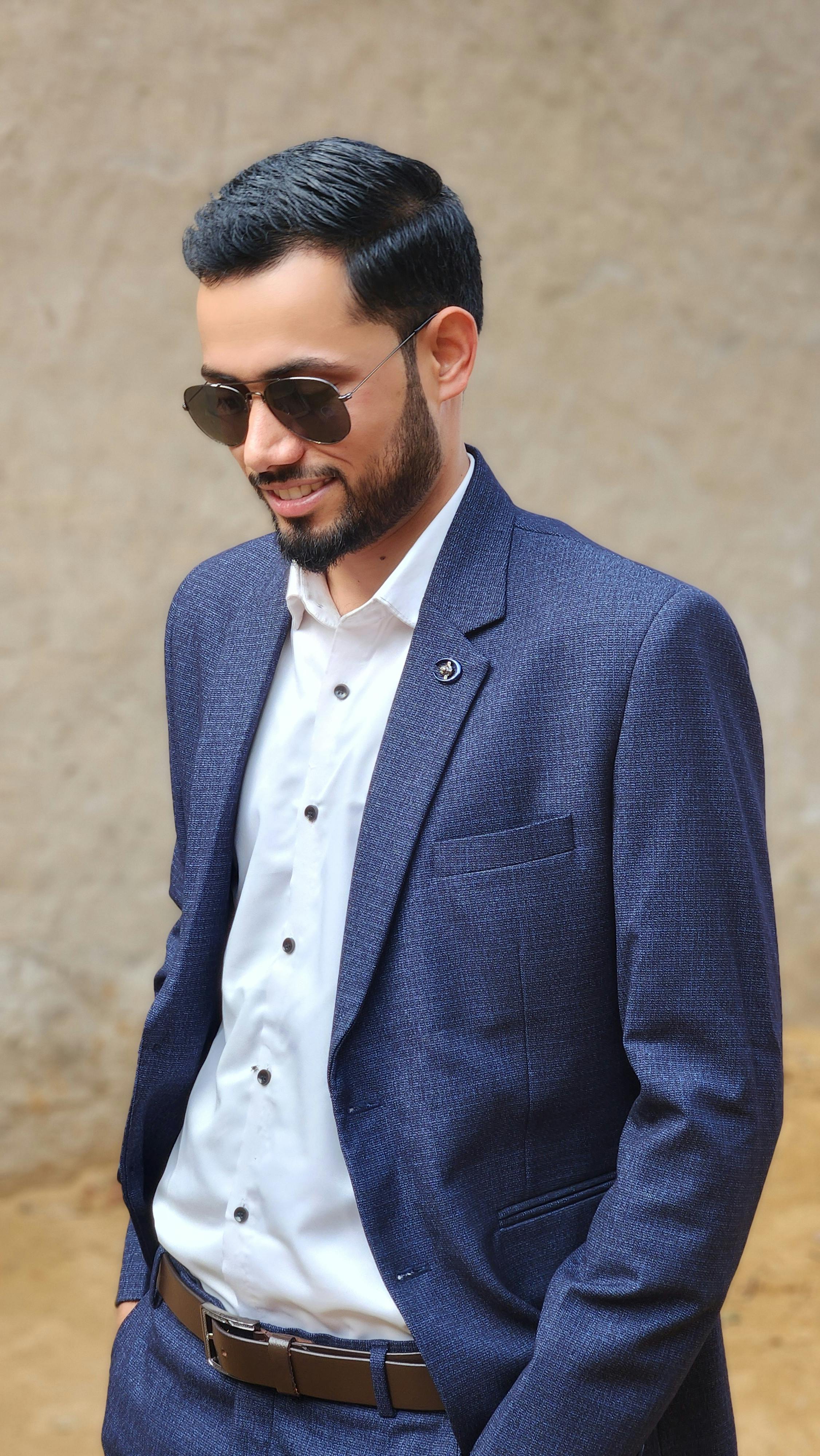 Confident businessman wearing a blue suit and sunglasses outdoors, smiling slightly.