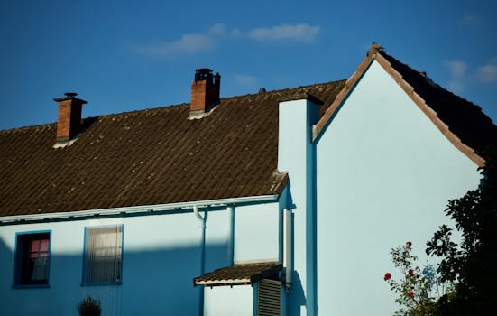 A picturesque blue house with a brown roof and chimney against a bright blue sky.