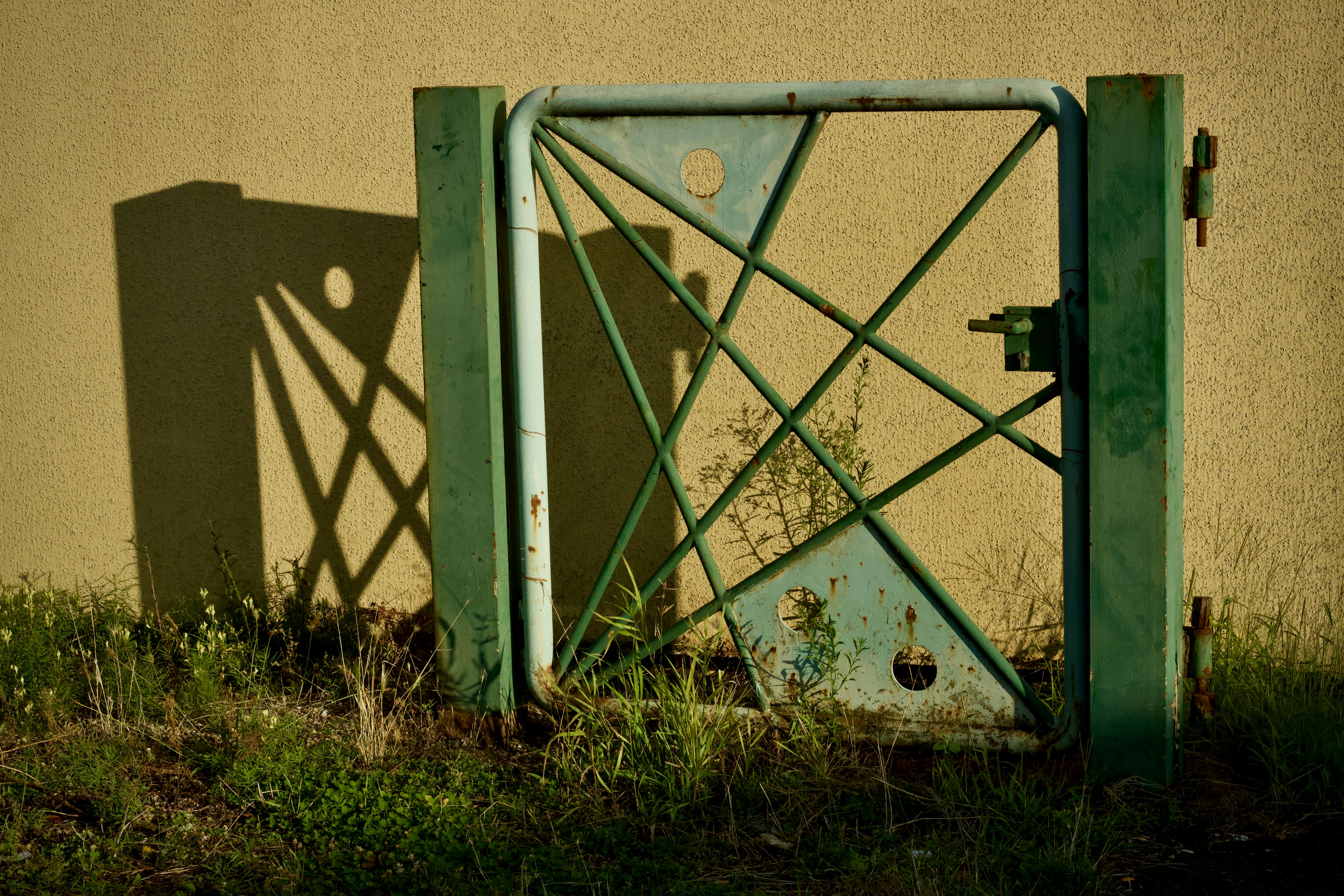 Free A rustic green metal gate casting a shadow on a sunlit wall with overgrown grass. Stock Photo