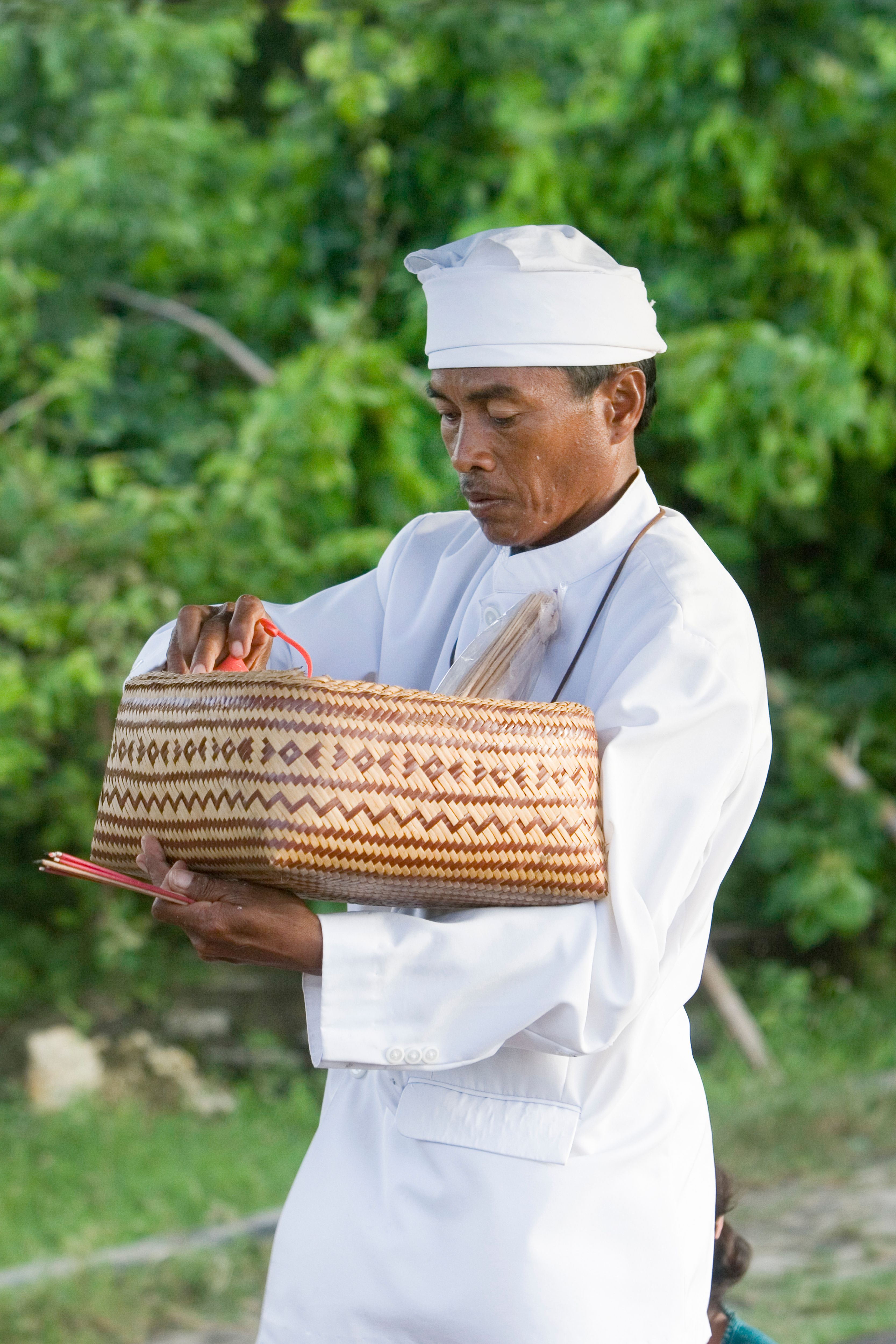 An adult man in ceremonial white clothing holding a traditional woven basket outdoors. - Fotografer Sidoarjo